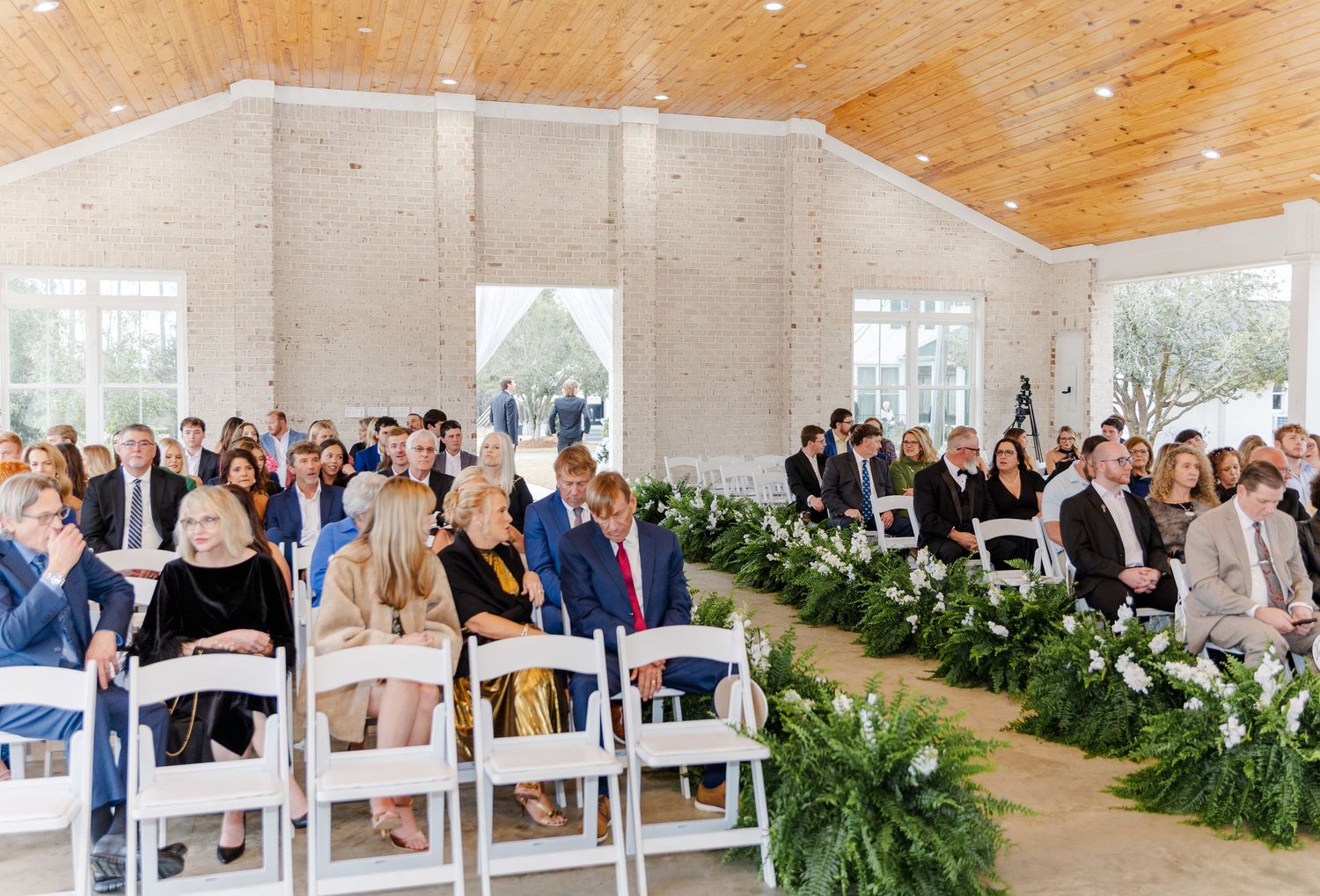 Guests seated in white chairs inside bright ceremony hall with greenery lining the aisle.