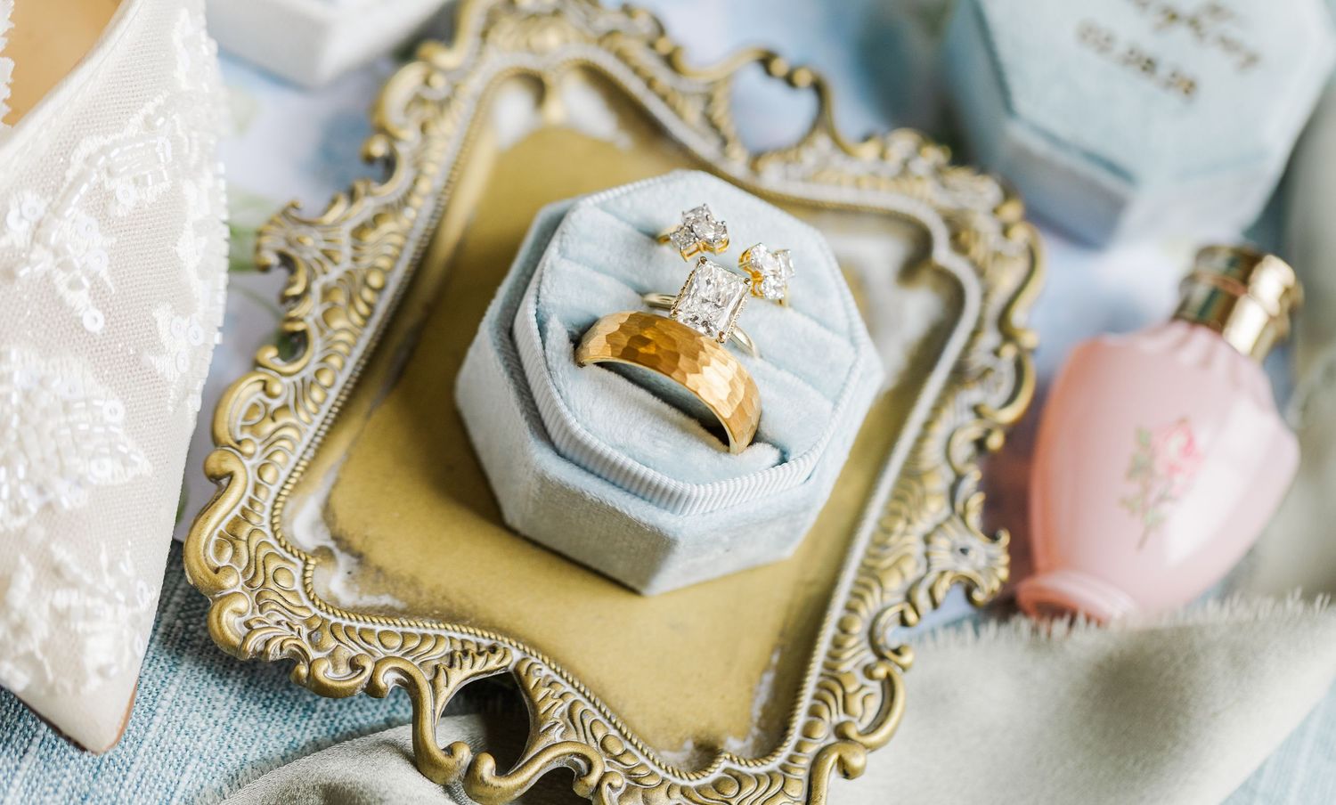 Gold diamond engagement ring displayed in a gray velvet ring box on an ornate gold tray with perfume bottles.