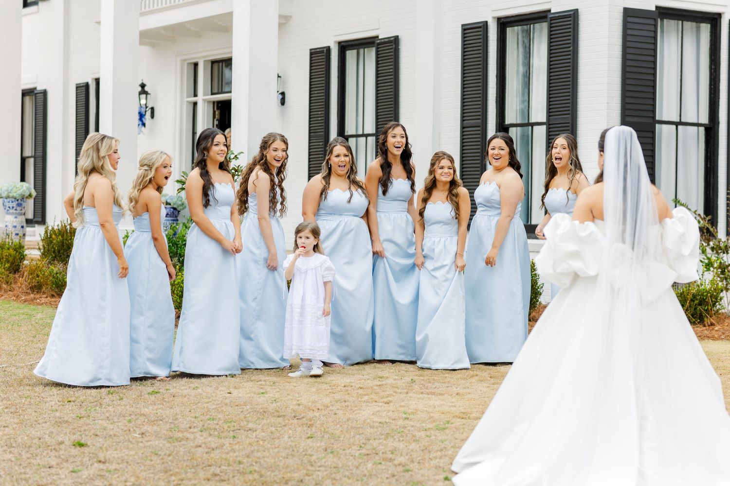 Bridesmaids in powder blue dresses react with joy during bride's first look reveal at outdoor wedding.