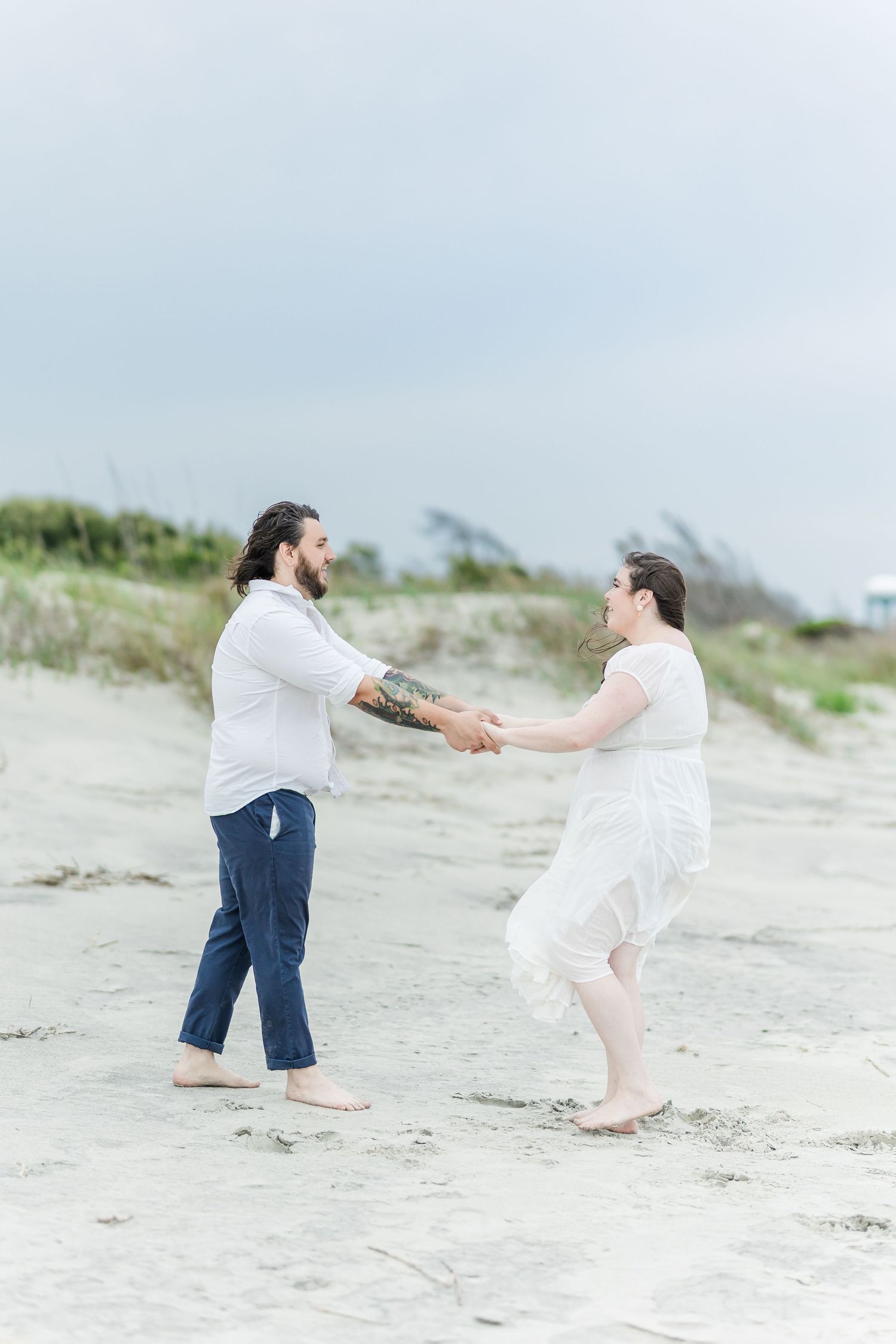 A playful couple dances together on a breezy beach wearing white and blue casual attire.