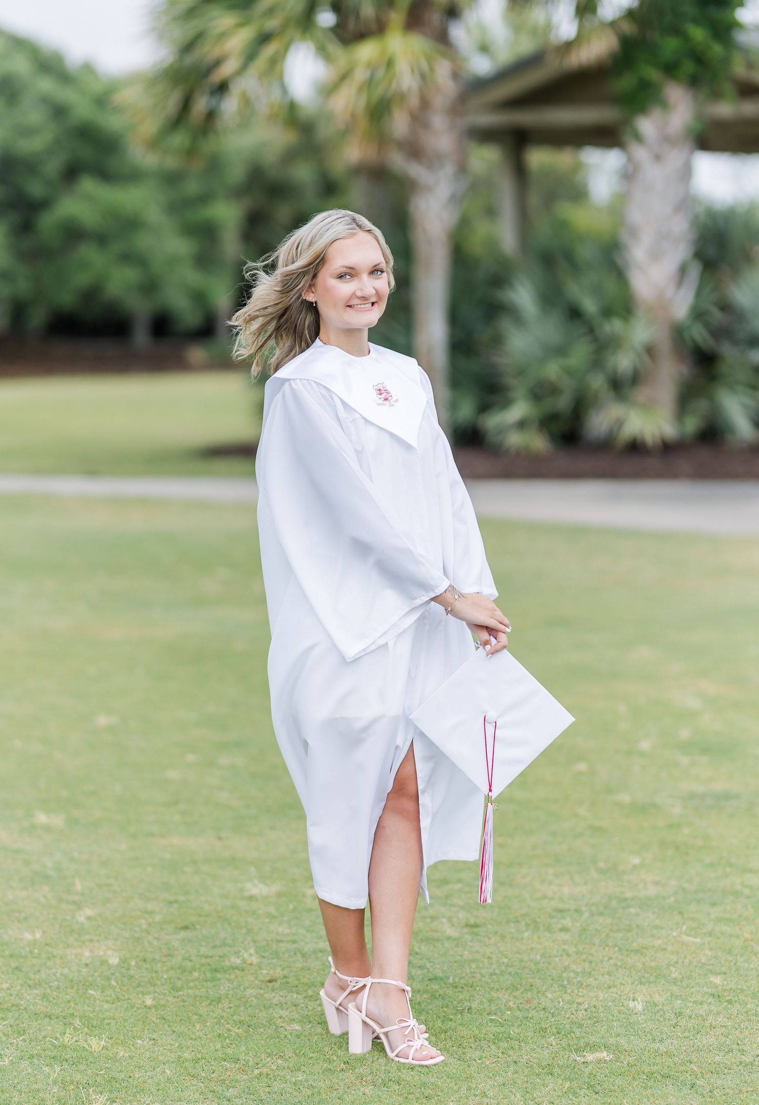 A graduate poses in white regalia against a backdrop of palm trees and green landscaping.