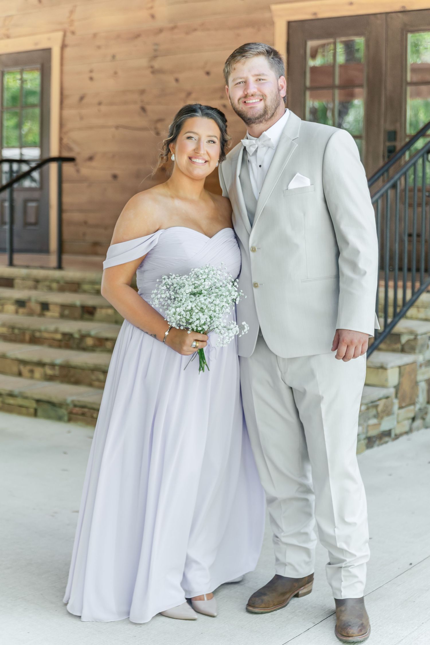 Couple in light formal wear smile together on wooden porch with bride holding baby's breath bouquet.