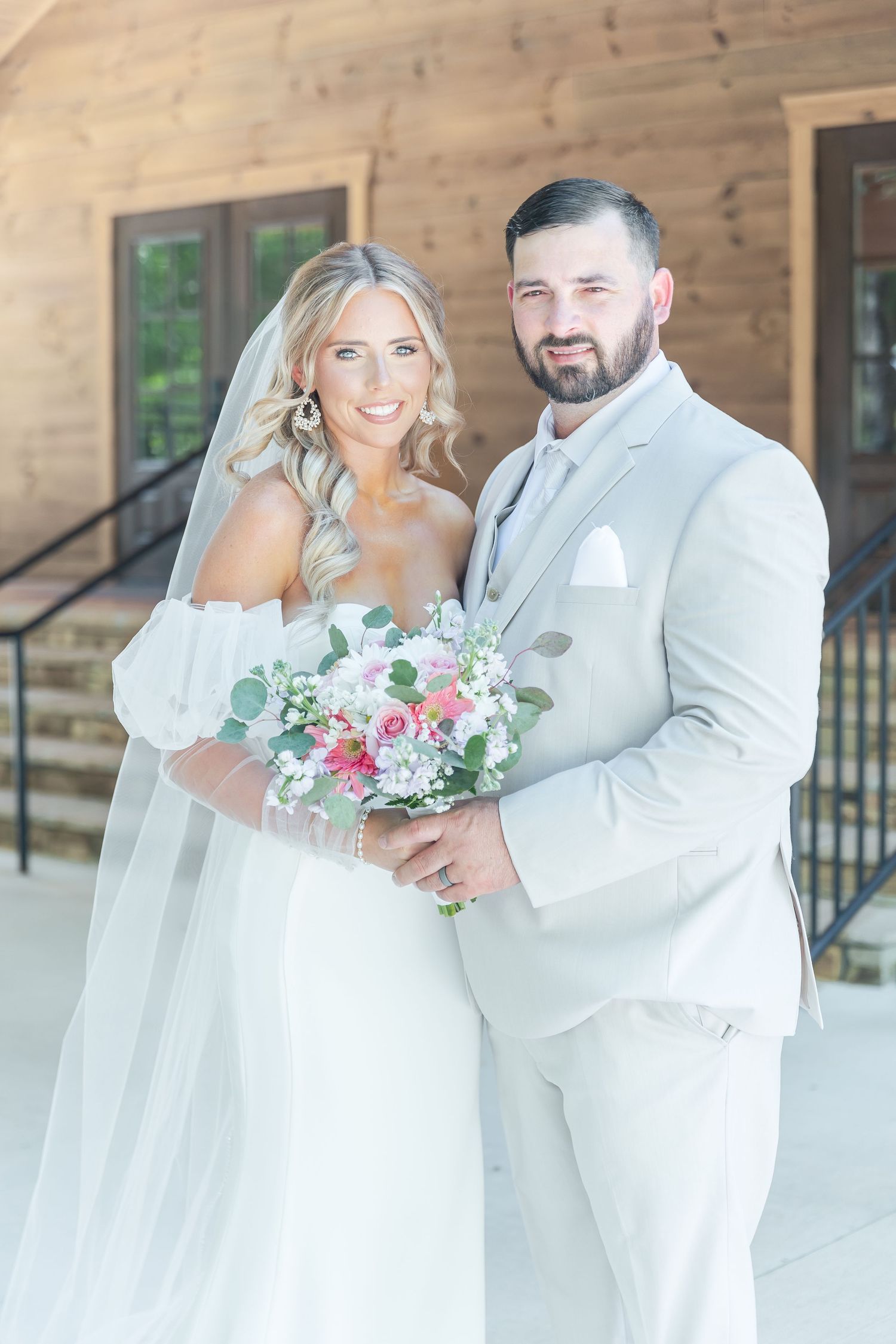 Wedding couple in white attire poses outdoors with bridal bouquet featuring pink and white flowers with greenery.