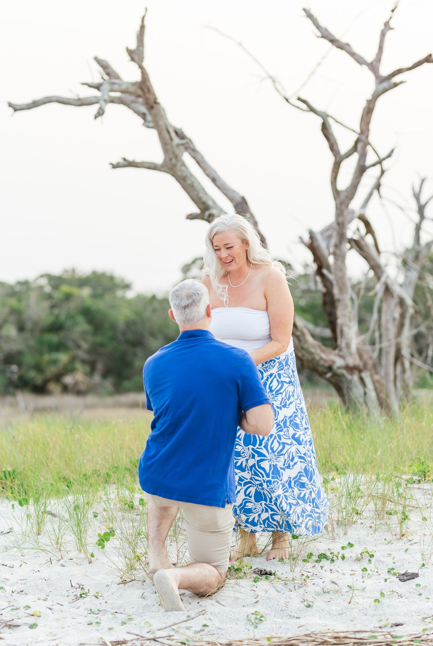 A heartwarming proposal scene with the couple standing near driftwood on a sandy beach.