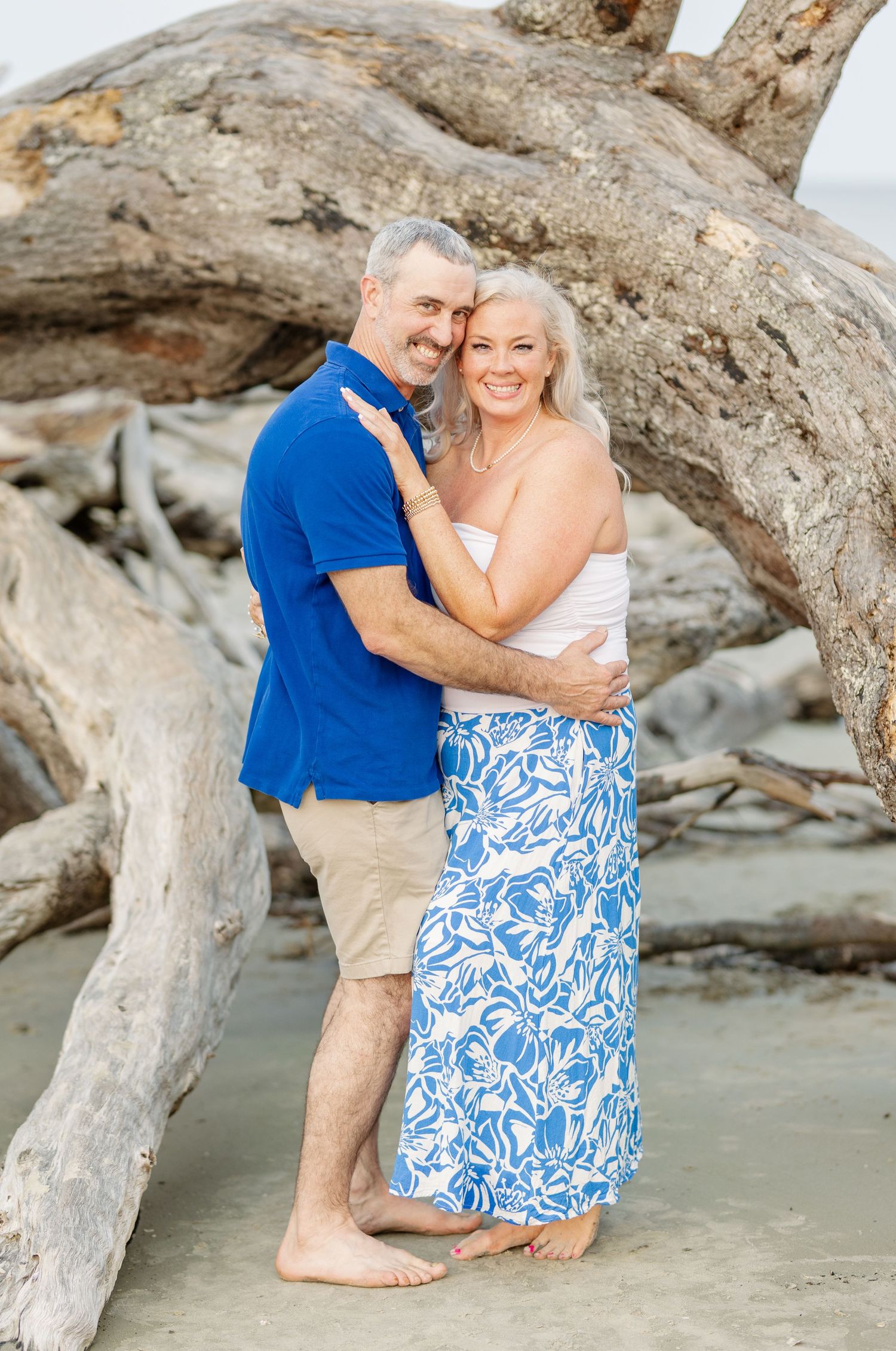 A couple embraces on a beach with driftwood, wearing blue and white coordinated outfits.