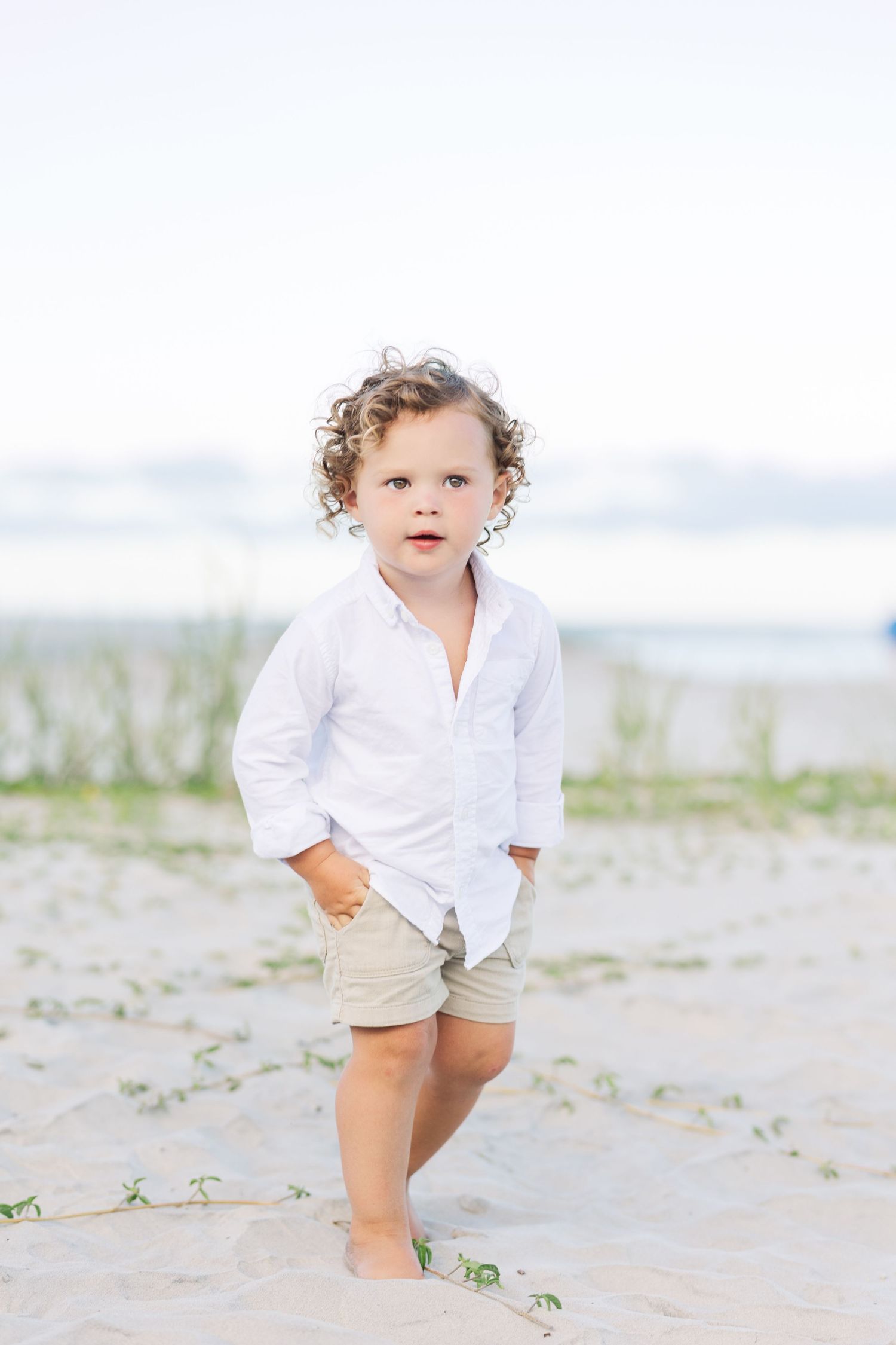 A toddler in a white shirt and khaki shorts poses on the beach with curly hair.