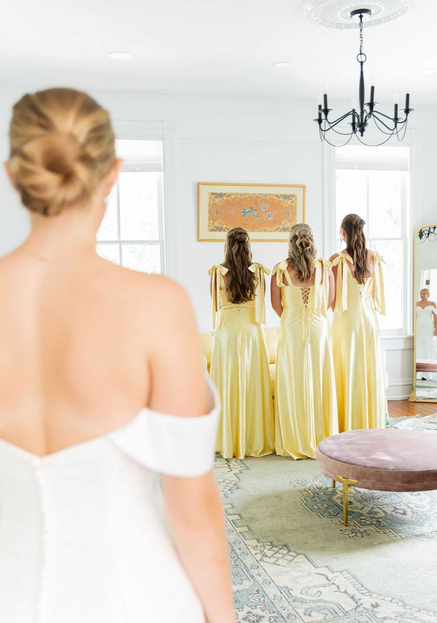 Bridesmaids in flowing pale yellow dresses stand together while bride in white gown watches from behind.