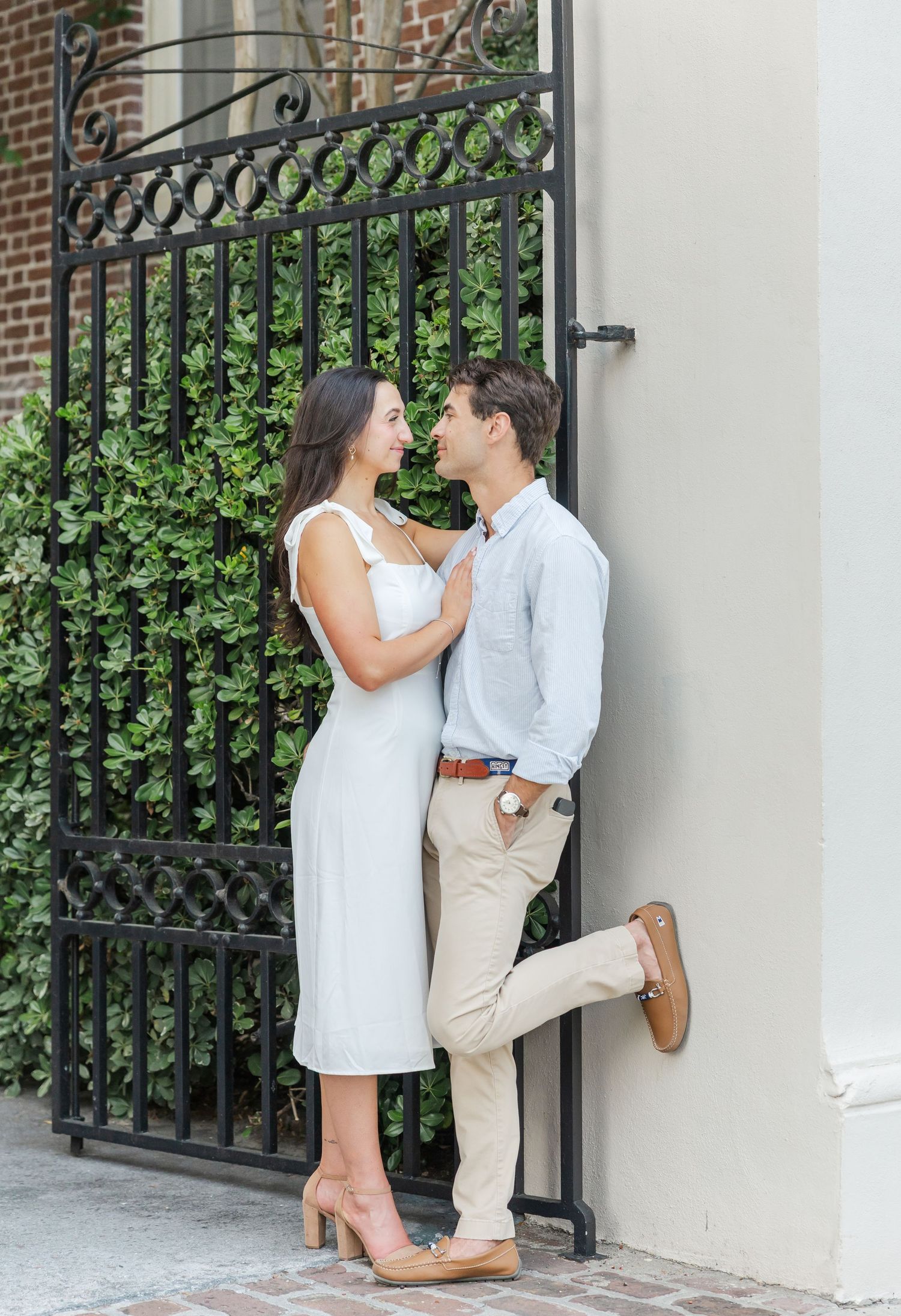 Couple gazing at each other smiling beside a black iron gate with lush green ivy backdrop.