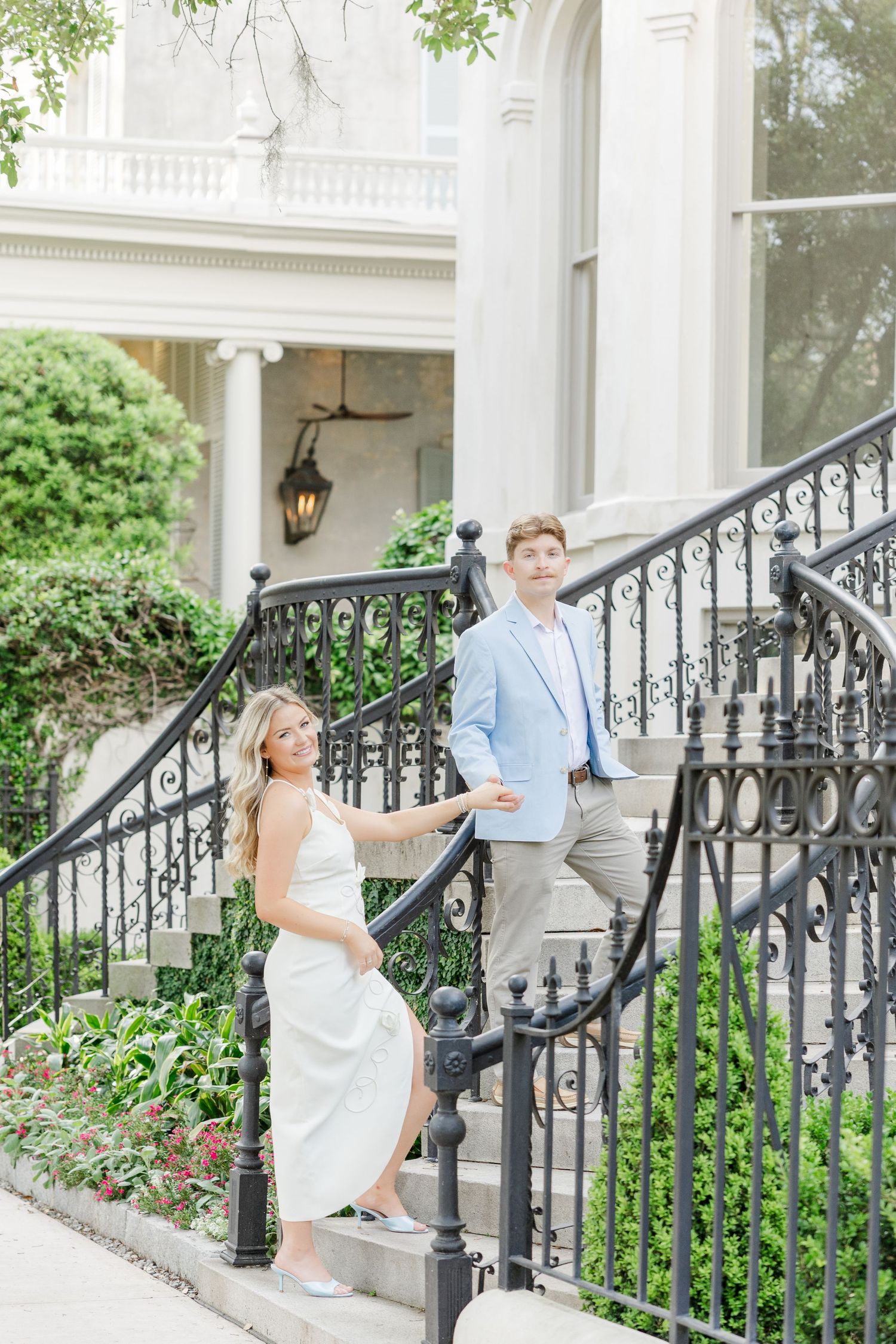 Couple posing on elegant iron staircase outside a white mansion, woman in white dress, man in light blue suit.