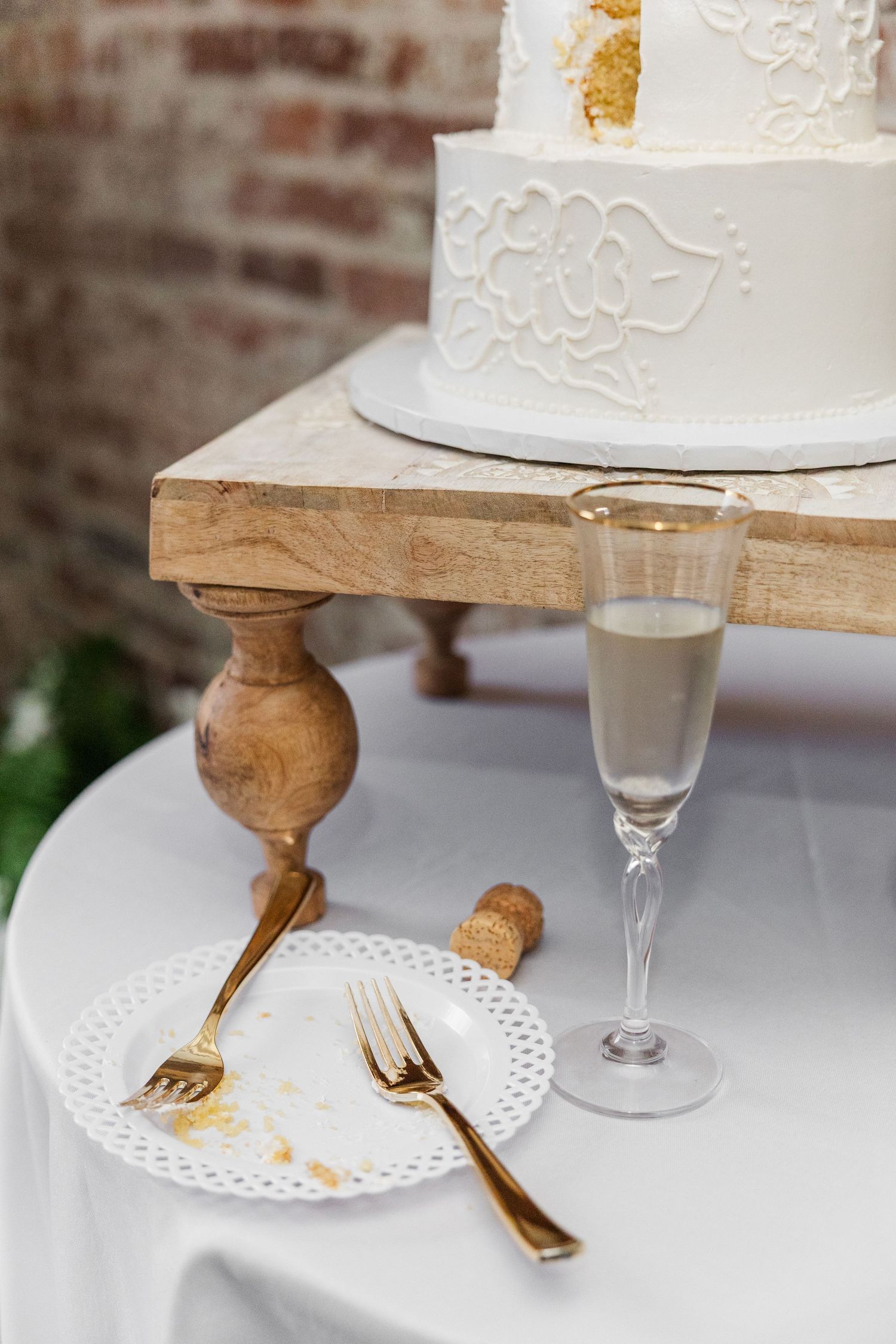 White wedding cake on rustic wood stand beside champagne flute and gold forks on white plate.