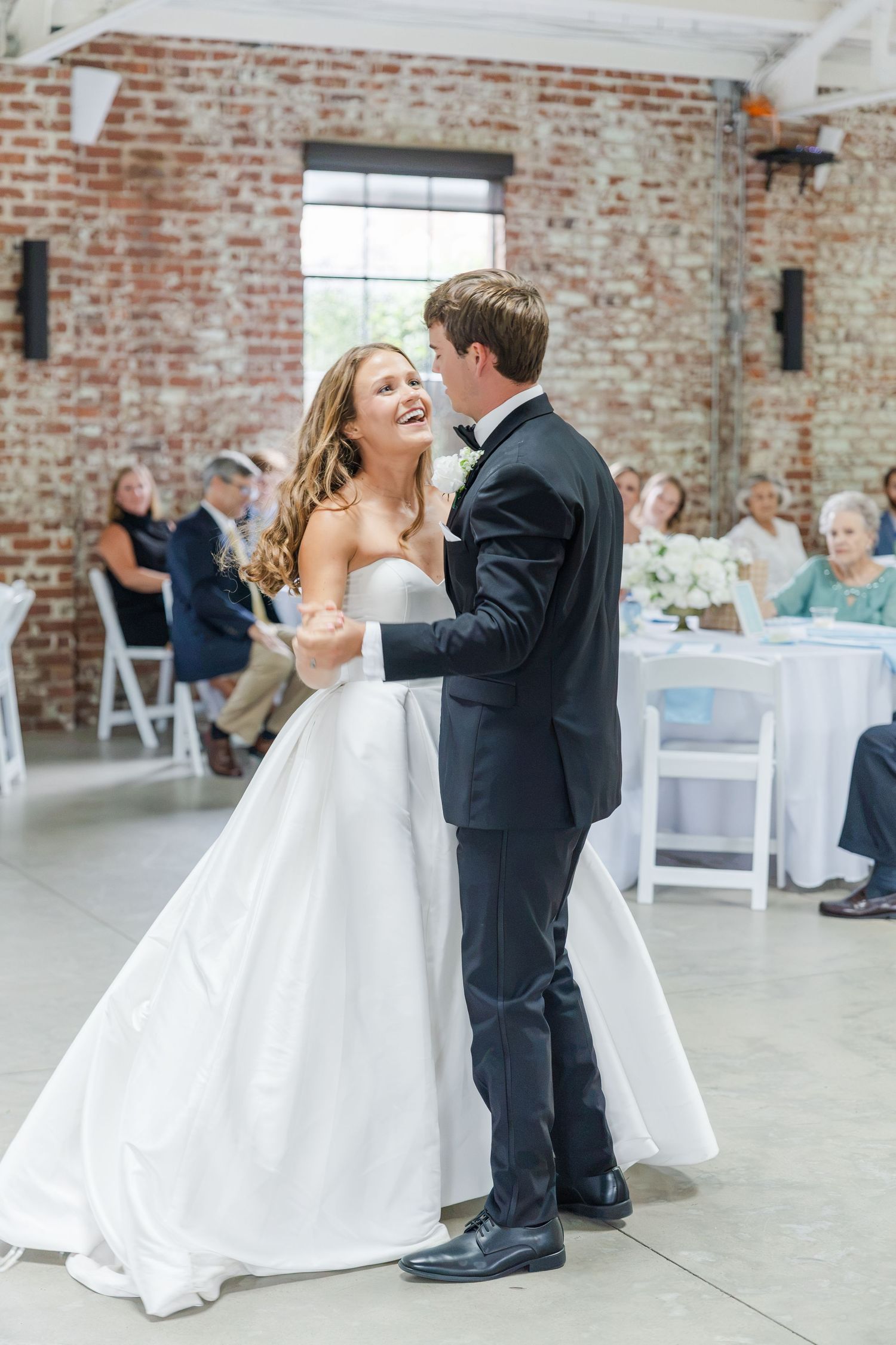 Bride and groom share their first dance, laughing joyfully, in elegant industrial venue with exposed brick walls.