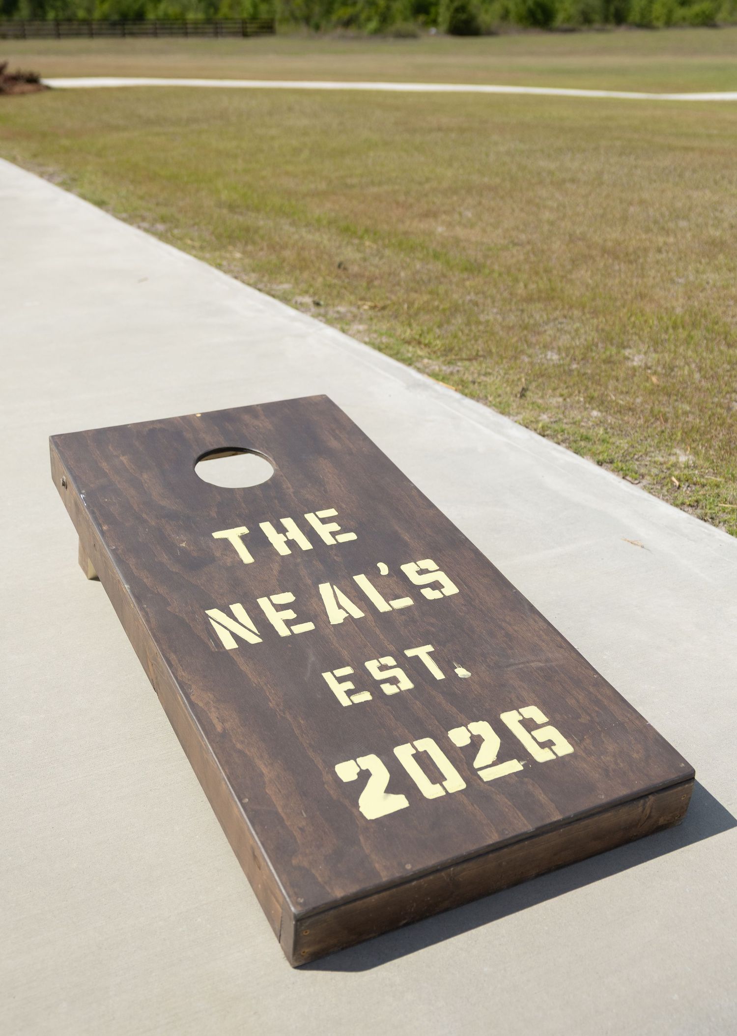 Custom personalized cornhole board reading The Neals Est. 2026 on a concrete path at outdoor wedding venue.