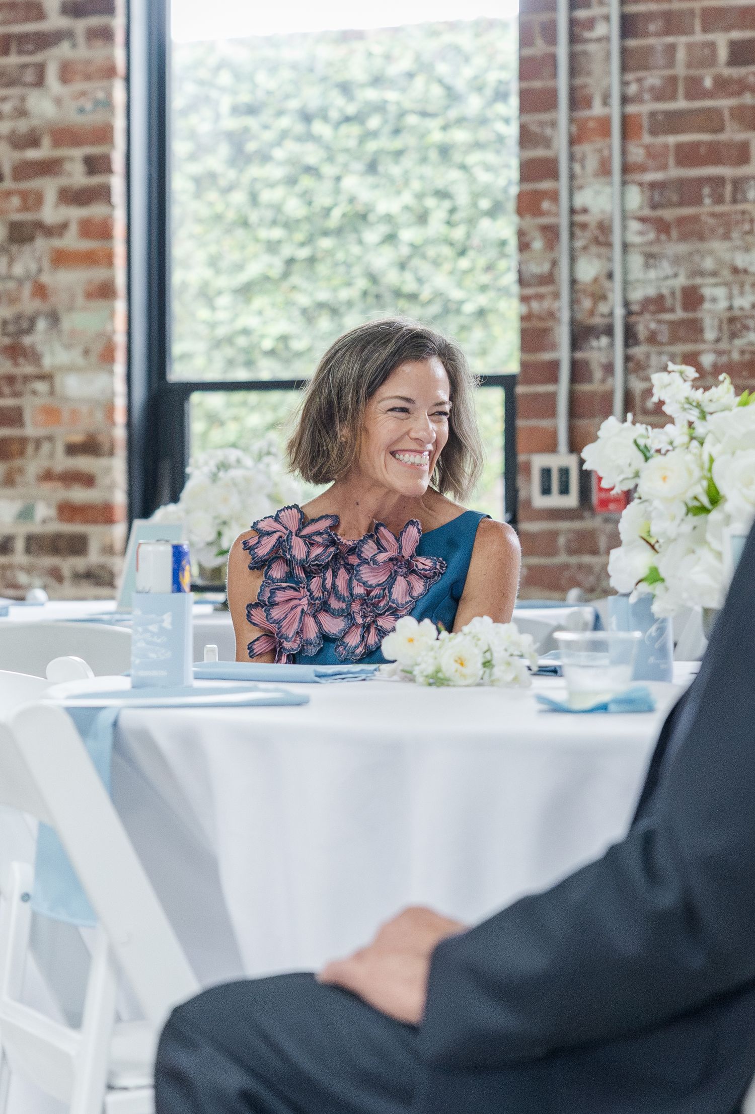 Smiling mother of the bride in teal floral dress seated at decorated wedding reception table.