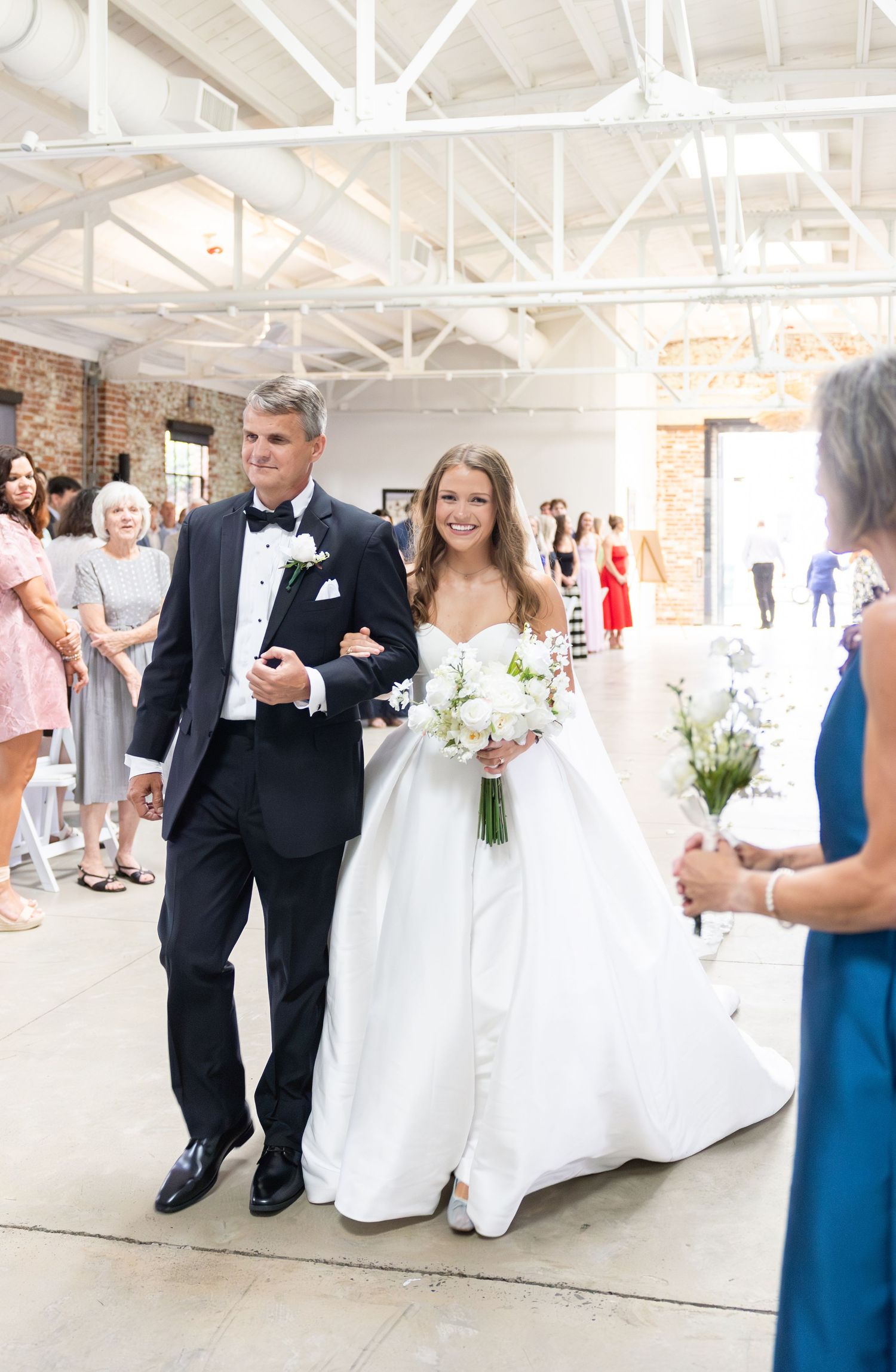 Father walks smiling bride in white ball gown down the aisle at elegant industrial wedding ceremony.