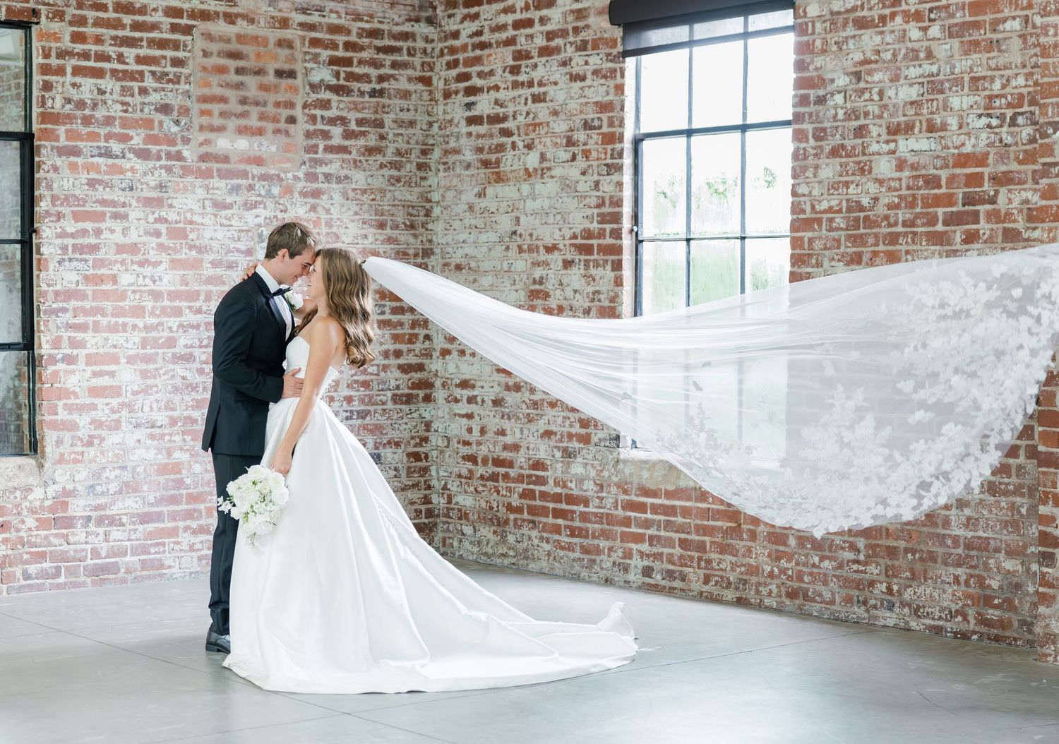 Bride and groom kissing as brides lace veil billows in wind inside brick-walled wedding venue.
