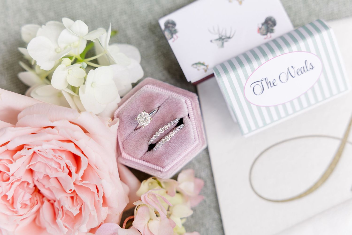 Pink velvet ring box with diamond engagement ring and wedding band beside blush roses and The Neals stationery.