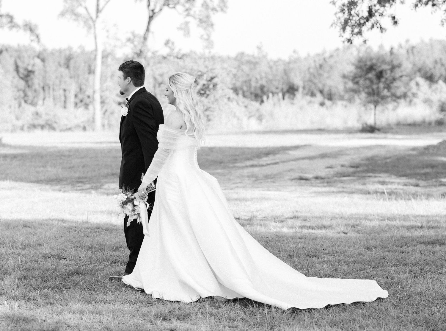 Black and white photo of bride and groom walking hand in hand across green lawn together.