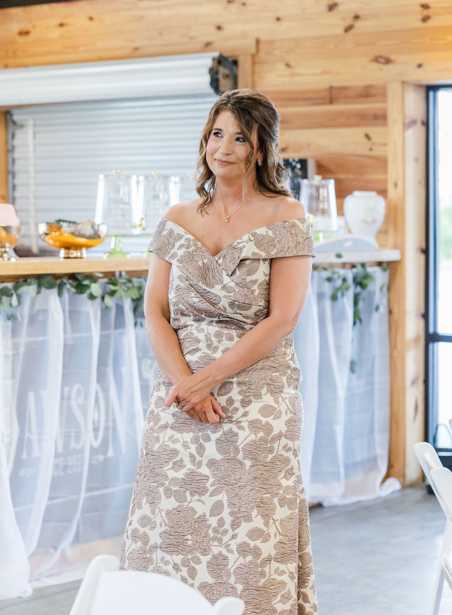 Elegant woman in off-shoulder floral gown stands smiling inside rustic barn wedding venue.