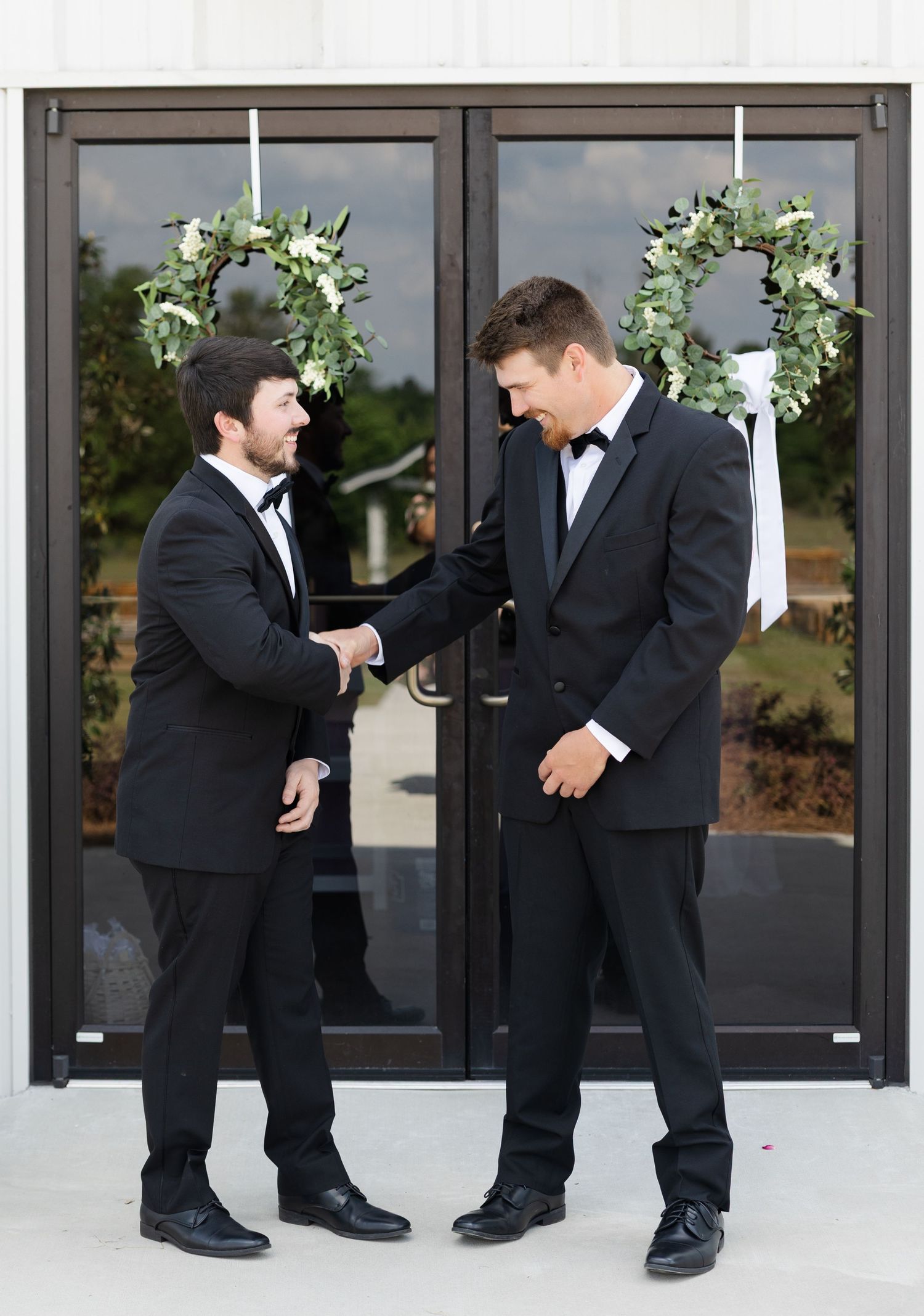 Two groomsmen in black tuxedos share a moment outside decorated church doors with greenery wreaths.