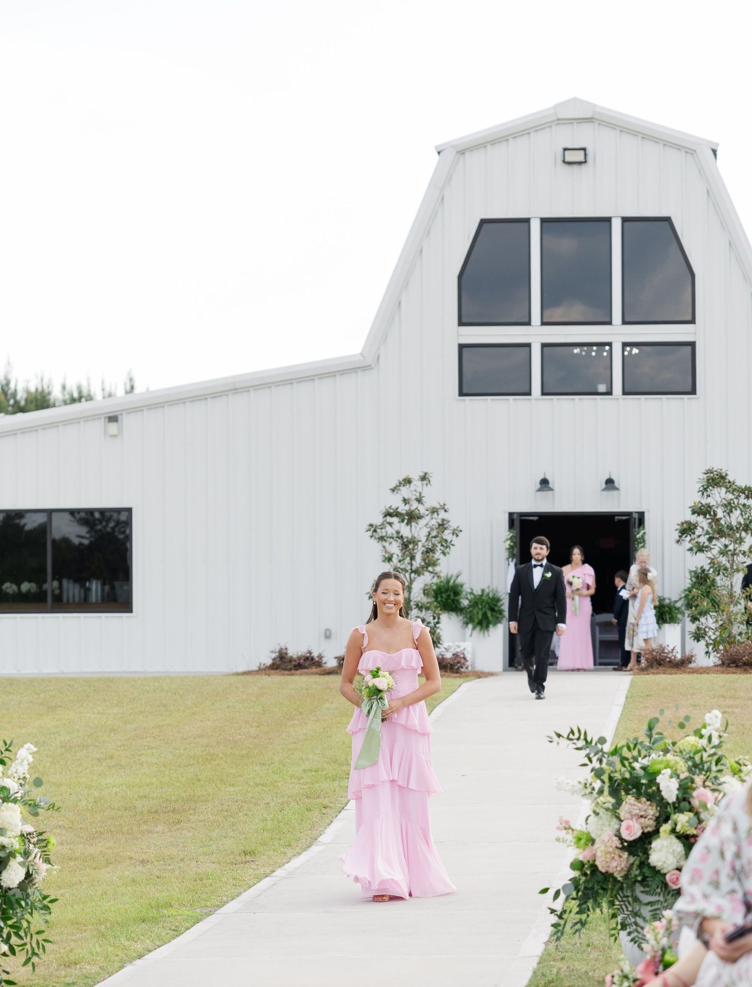 Bridesmaid in pink gown walks outdoor aisle toward white barn wedding venue.