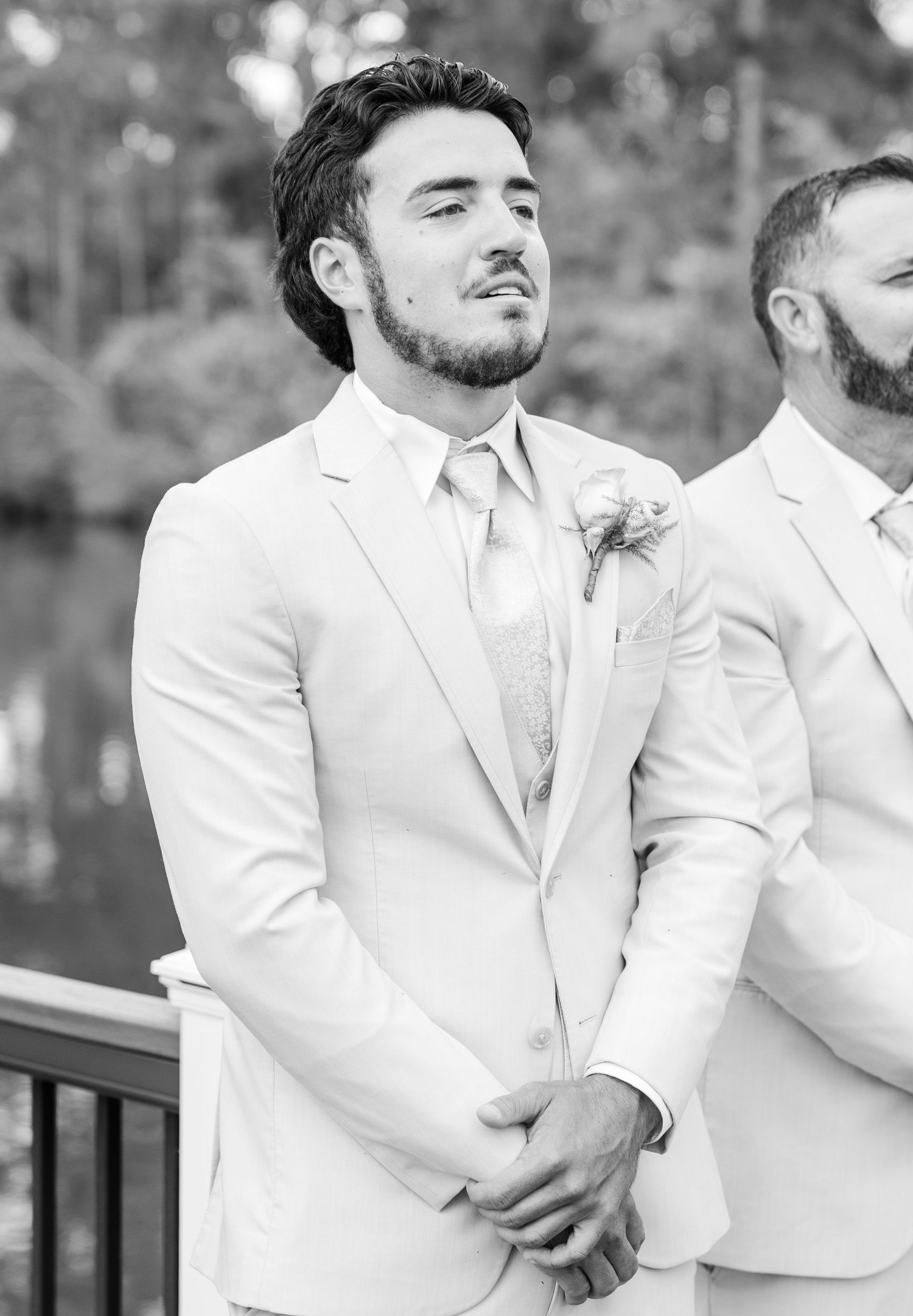Emotional groom in light suit with boutonniere awaits bride during outdoor wedding ceremony in black and white photo.