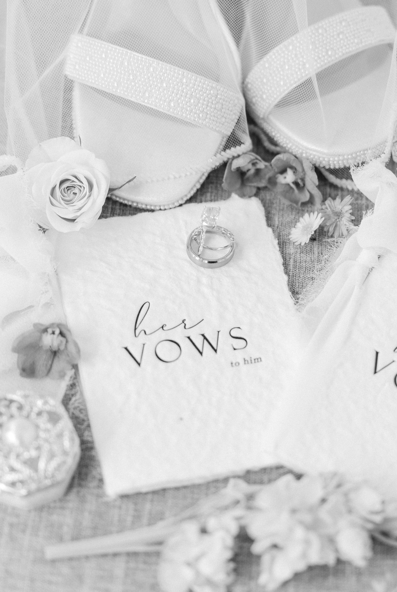 Black and white flat lay of bridal vows book, wedding rings, pearl heels, roses, and perfume bottle.
