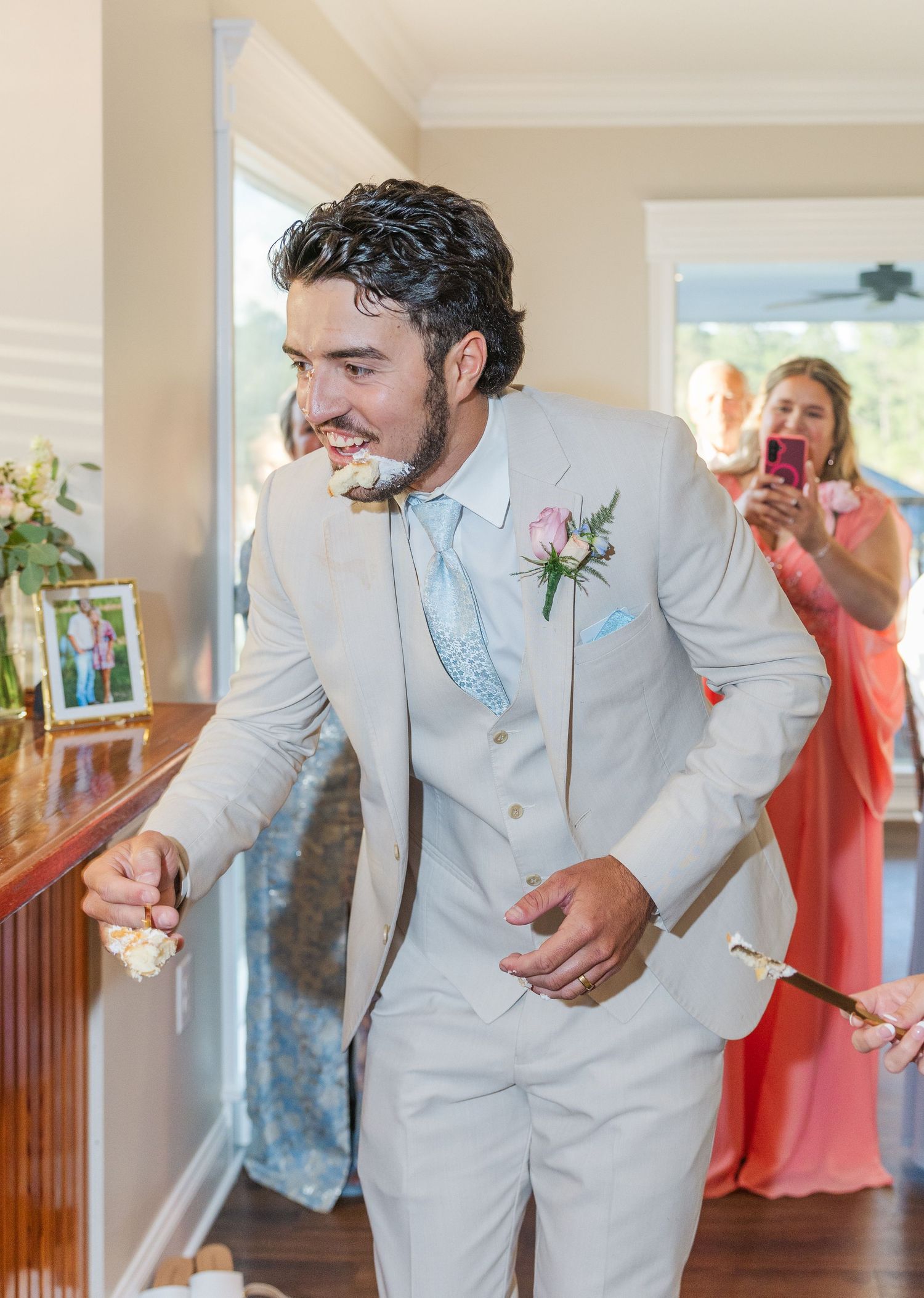 Groom in beige suit laughs while eating wedding cake as guests capture moment on phones.