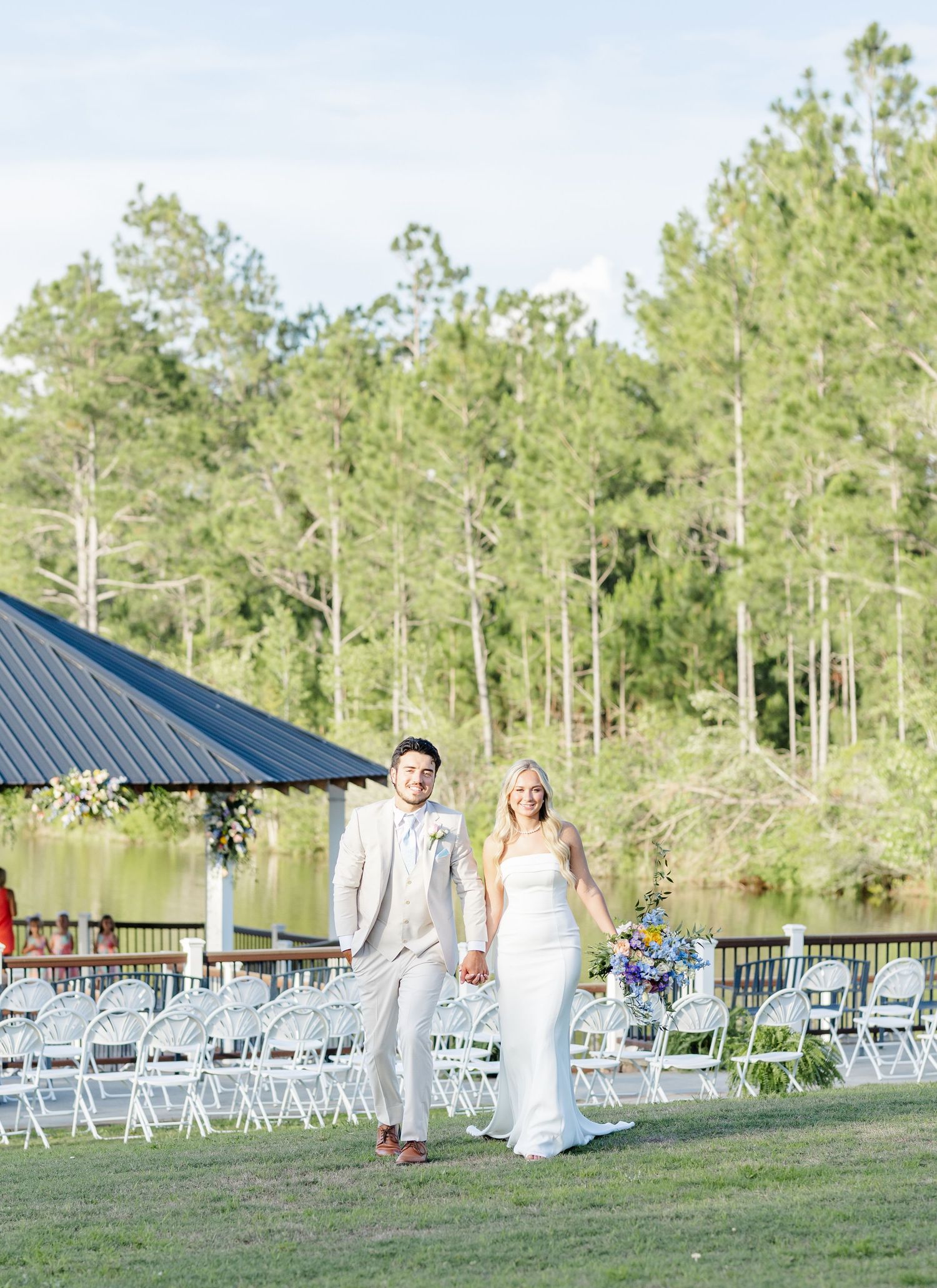 Newlyweds walking hand-in-hand down outdoor aisle with lake and pine trees in background.