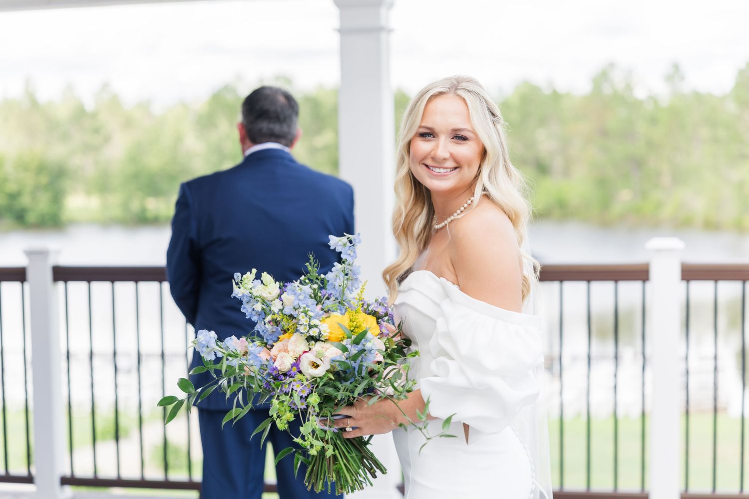 Smiling bride holding colorful blue and yellow bouquet during first look on outdoor venue balcony.