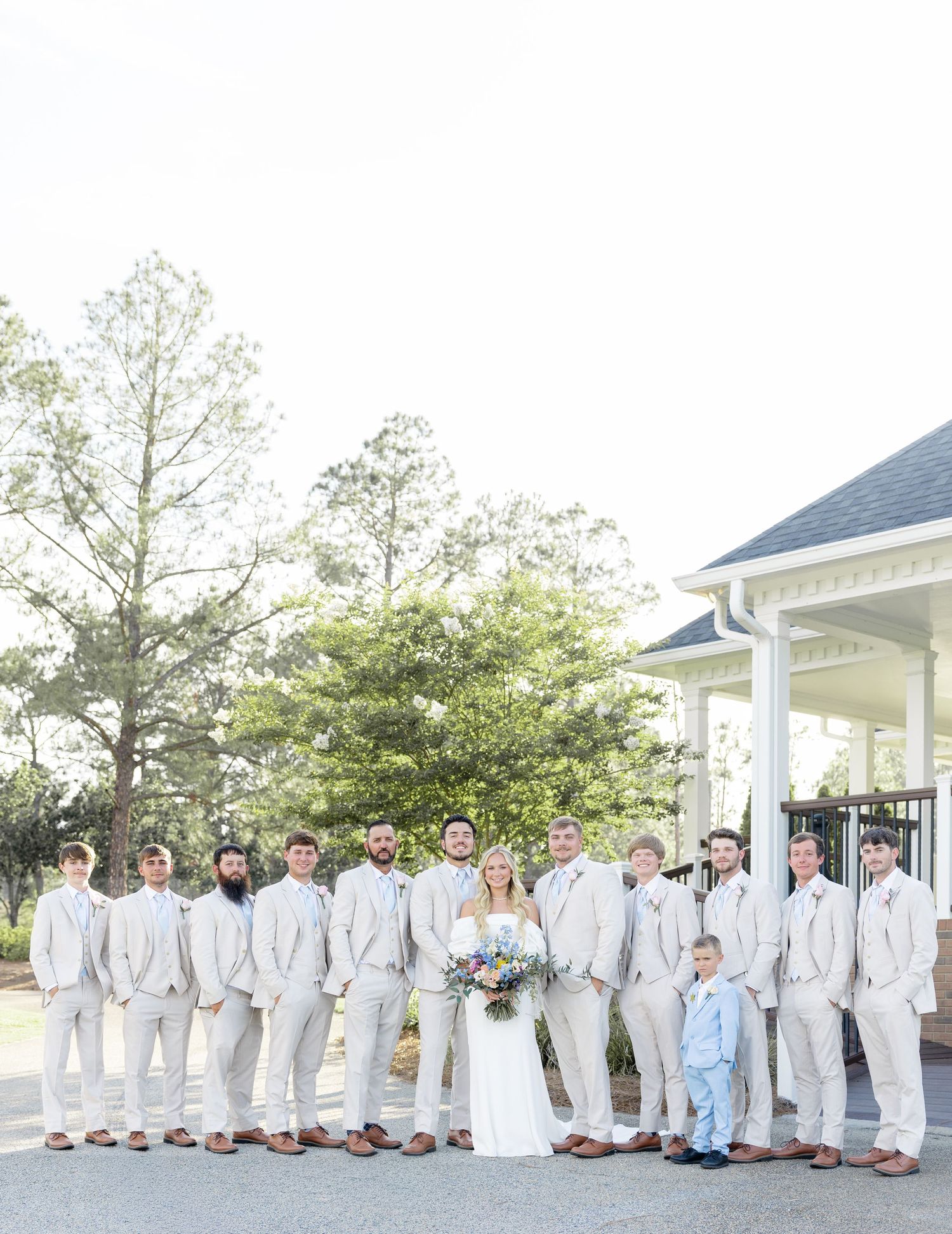Bride surrounded by large groomsmen party in matching light gray suits outside white wedding venue.