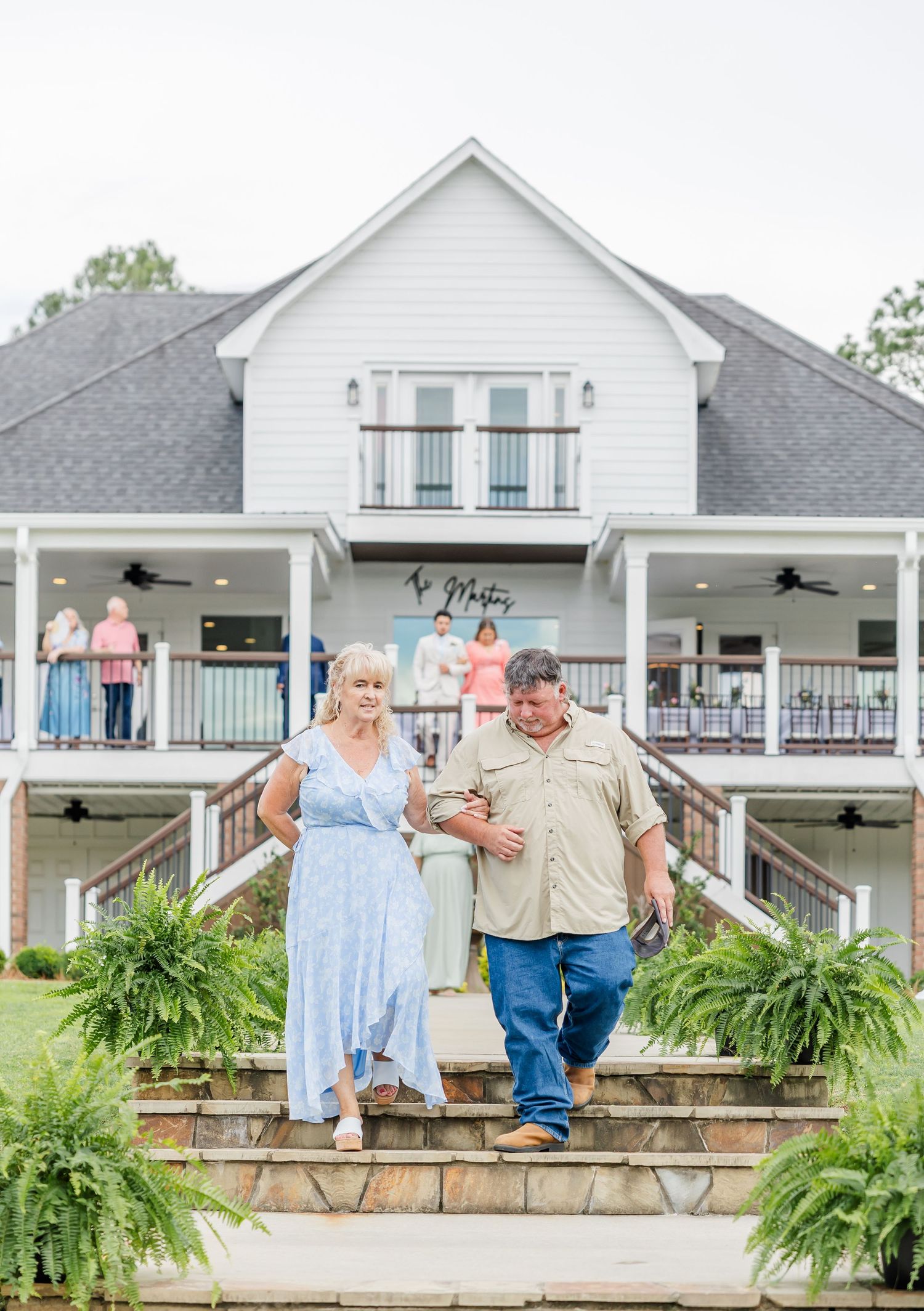 Couple walking down outdoor steps of white farmhouse wedding venue surrounded by lush greenery.