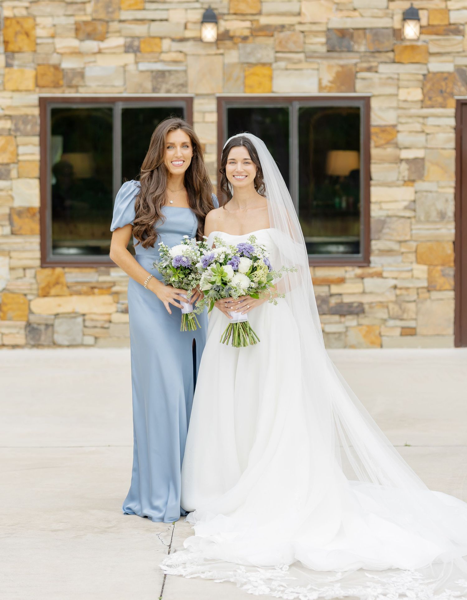 Wedding party poses against stone building exterior with elegant floral arrangements.