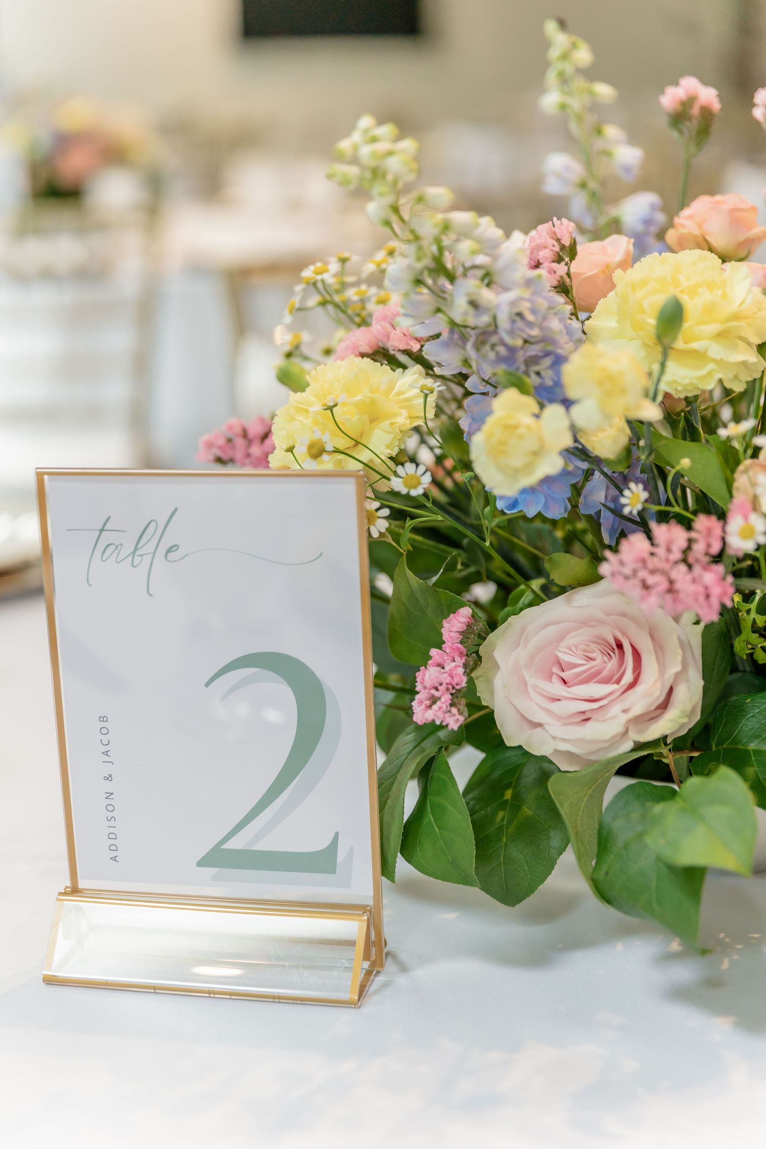 Pastel floral centerpiece with pink roses and yellow blooms alongside table number two in gold frame.