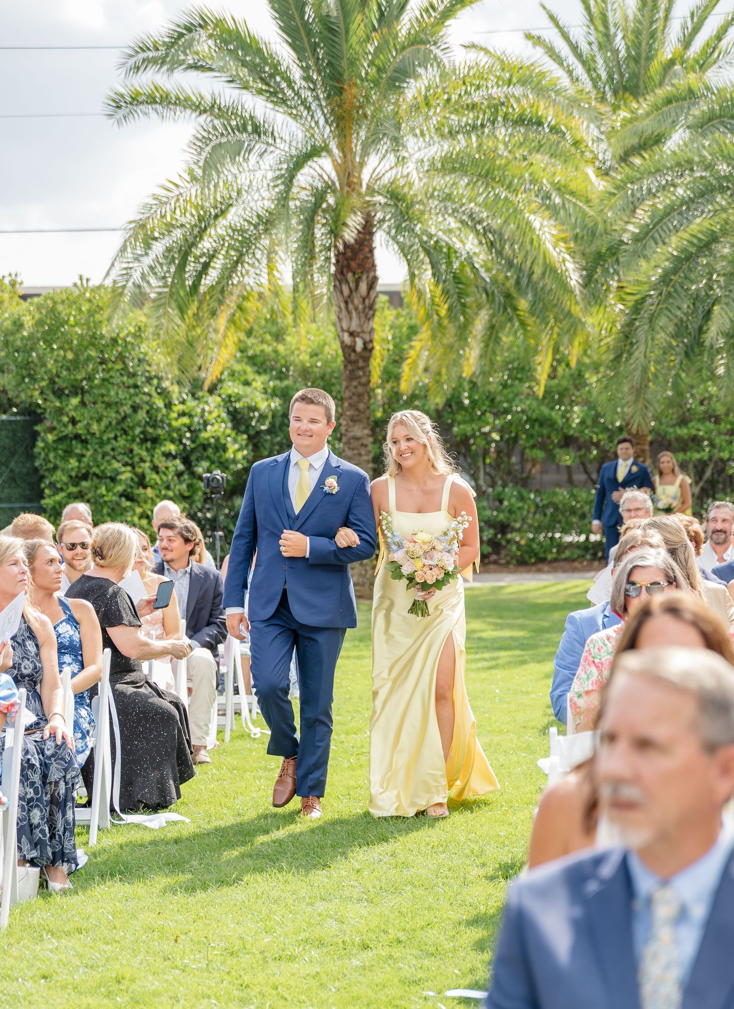 A wedding ceremony procession walks down a lush garden aisle lined with palm trees and seated guests.