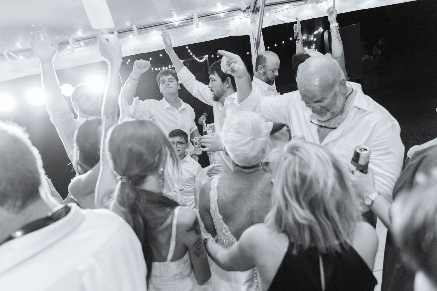 Wedding guests dancing and celebrating on a dance floor under white tent lighting.