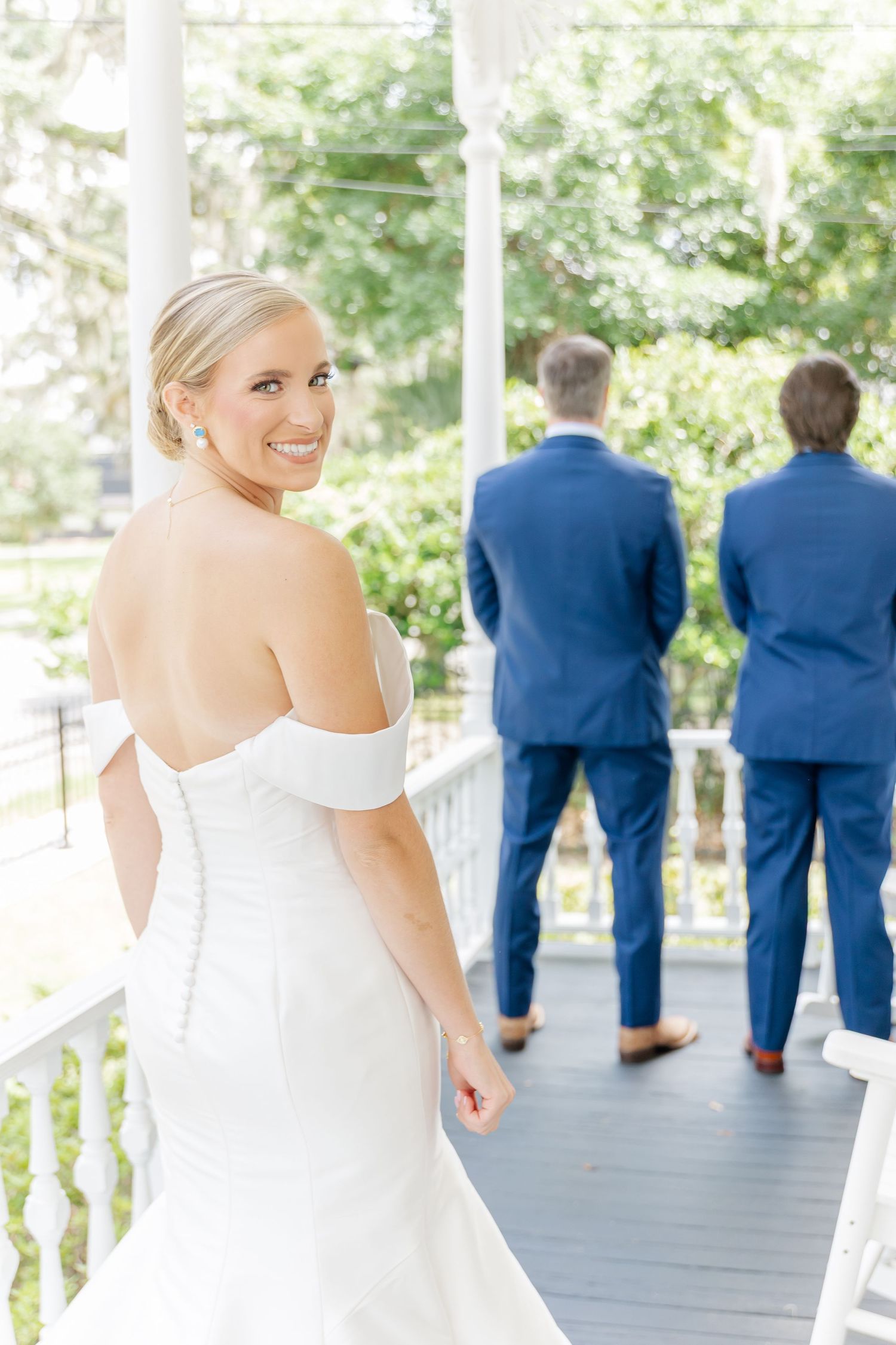 Wedding party members in navy suits and white dress stand on an outdoor porch.