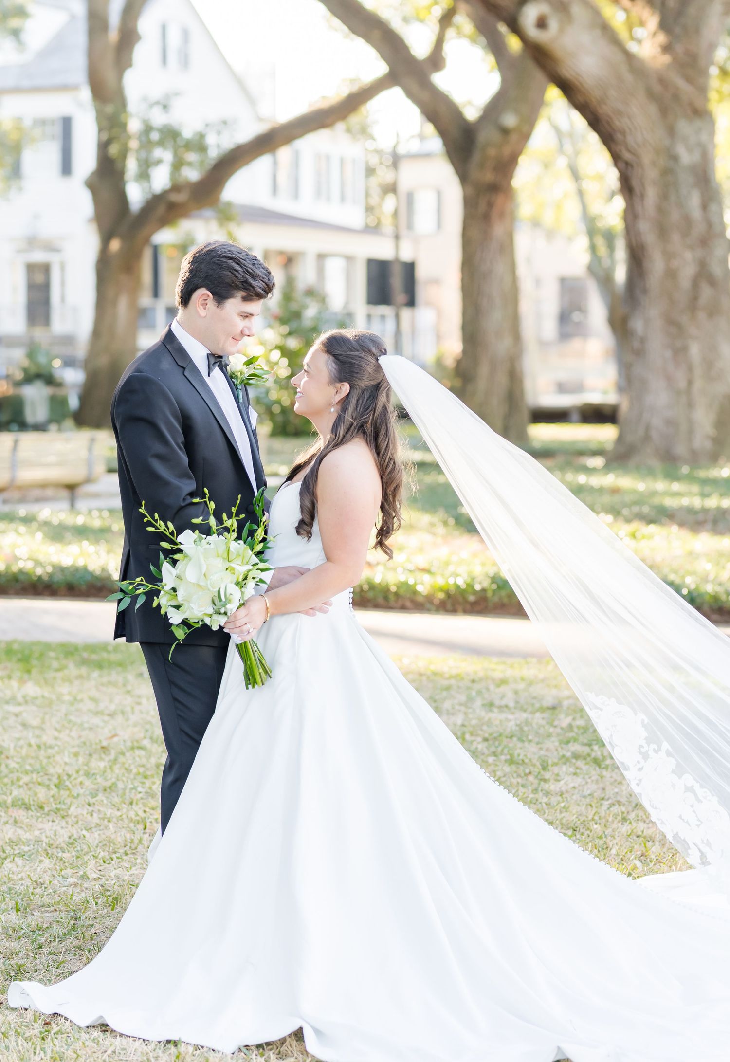 Bride in white ball gown with flowing veil and groom in black tuxedo share a romantic moment under oak trees.