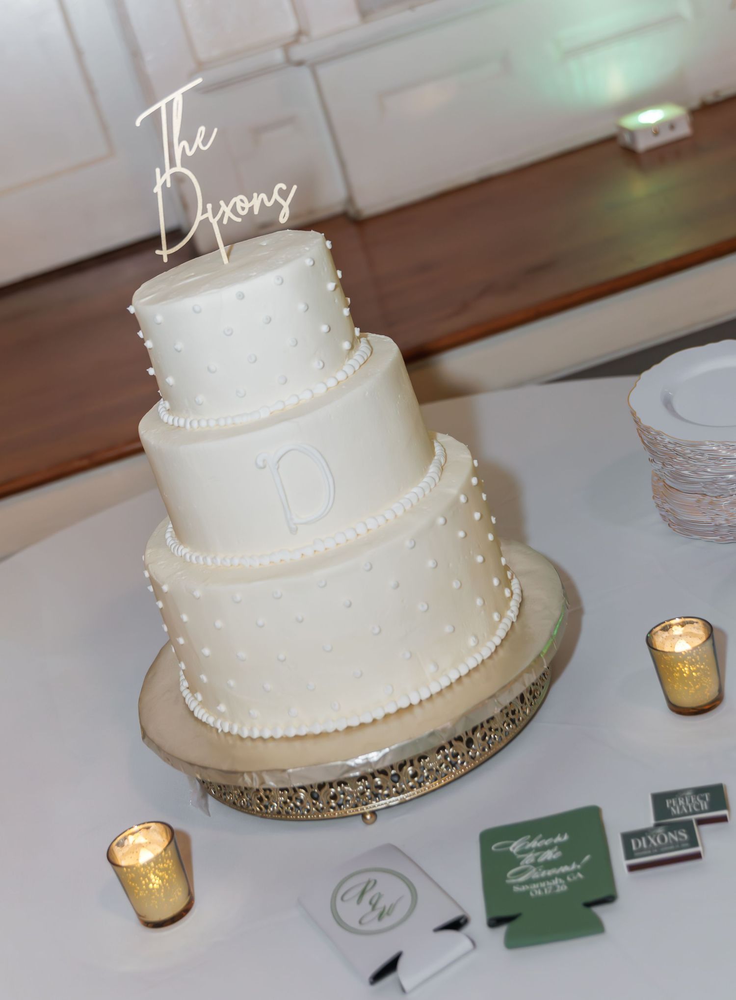 Three-tier white wedding cake with pearl details and The Dixons gold script topper on rhinestone cake stand.