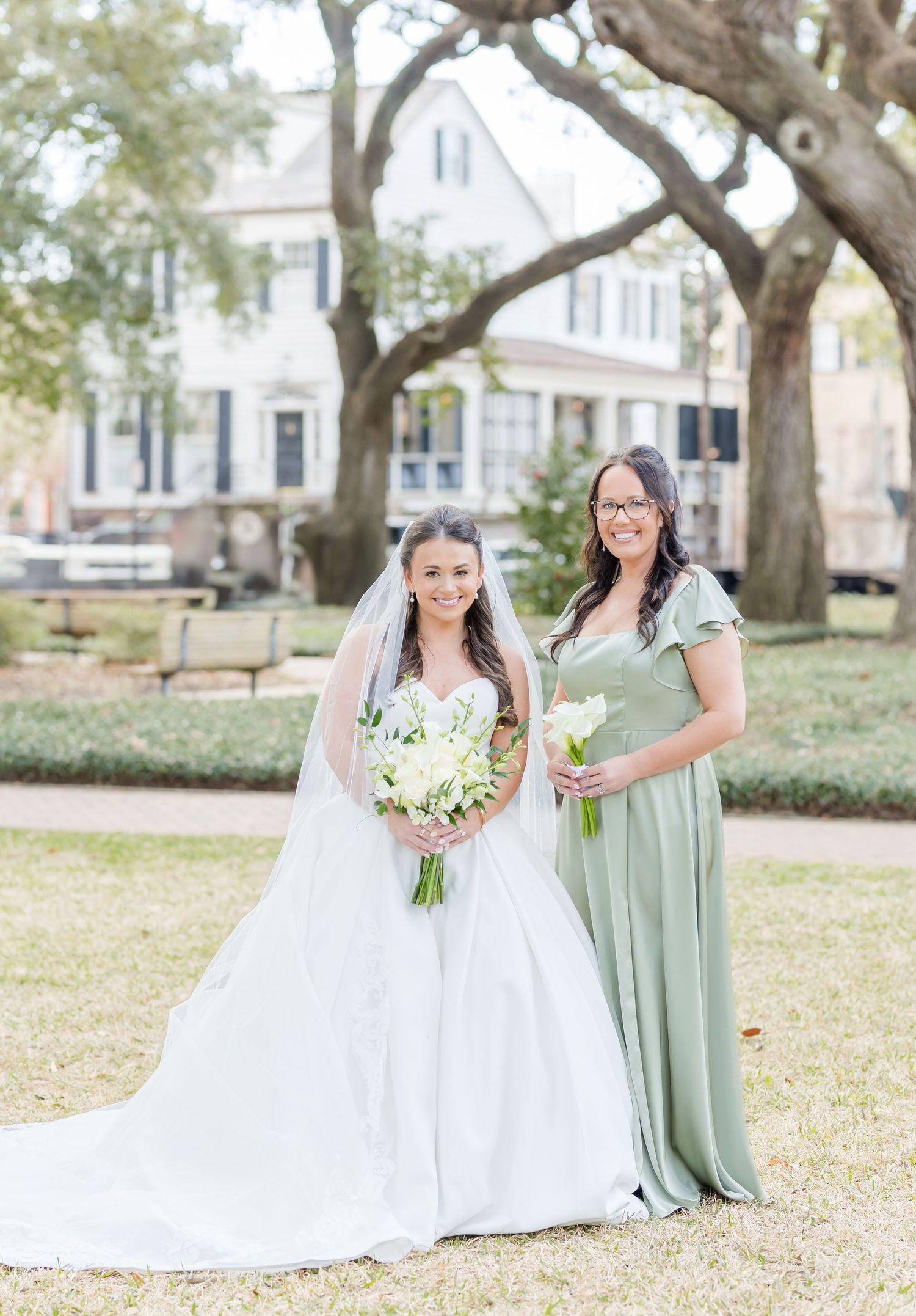 Bride in white gown with veil holds bouquet beside bridesmaid in sage green dress outdoors.