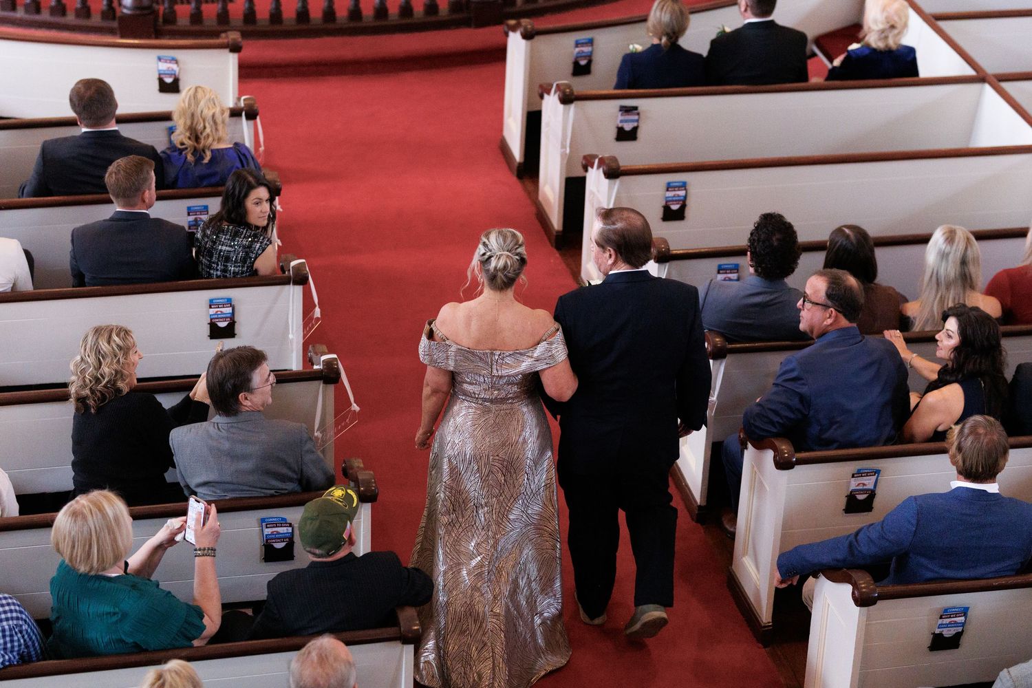 Aerial view of wedding guests seated in church pews, many holding phones, as a couple walks down the aisle.