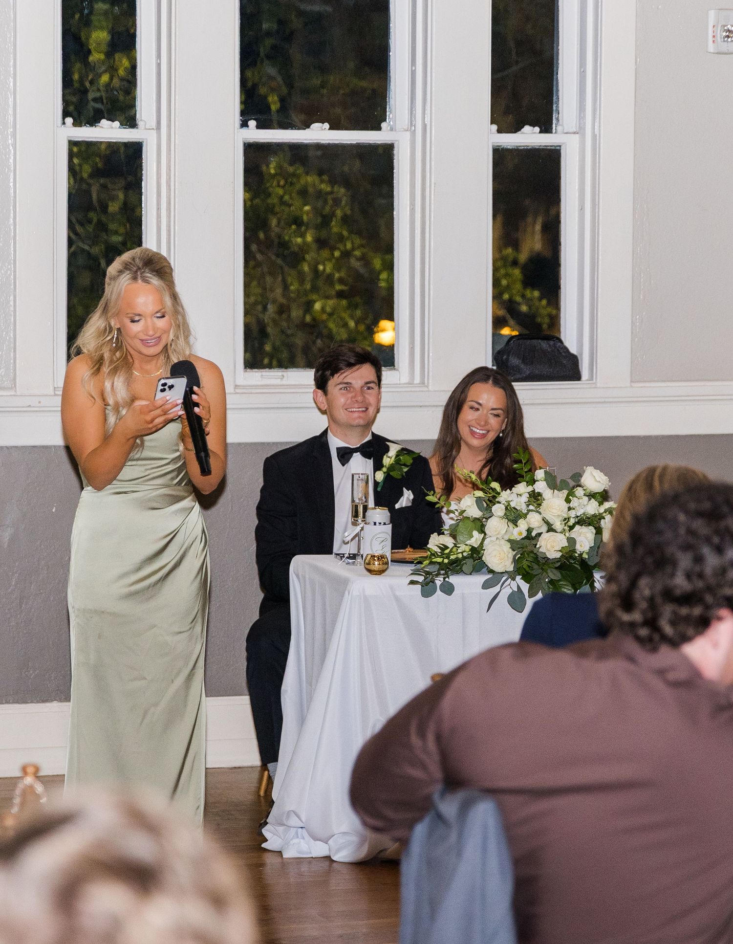 Bridesmaid gives a toast speech at wedding reception while bride and groom smile at sweetheart table.