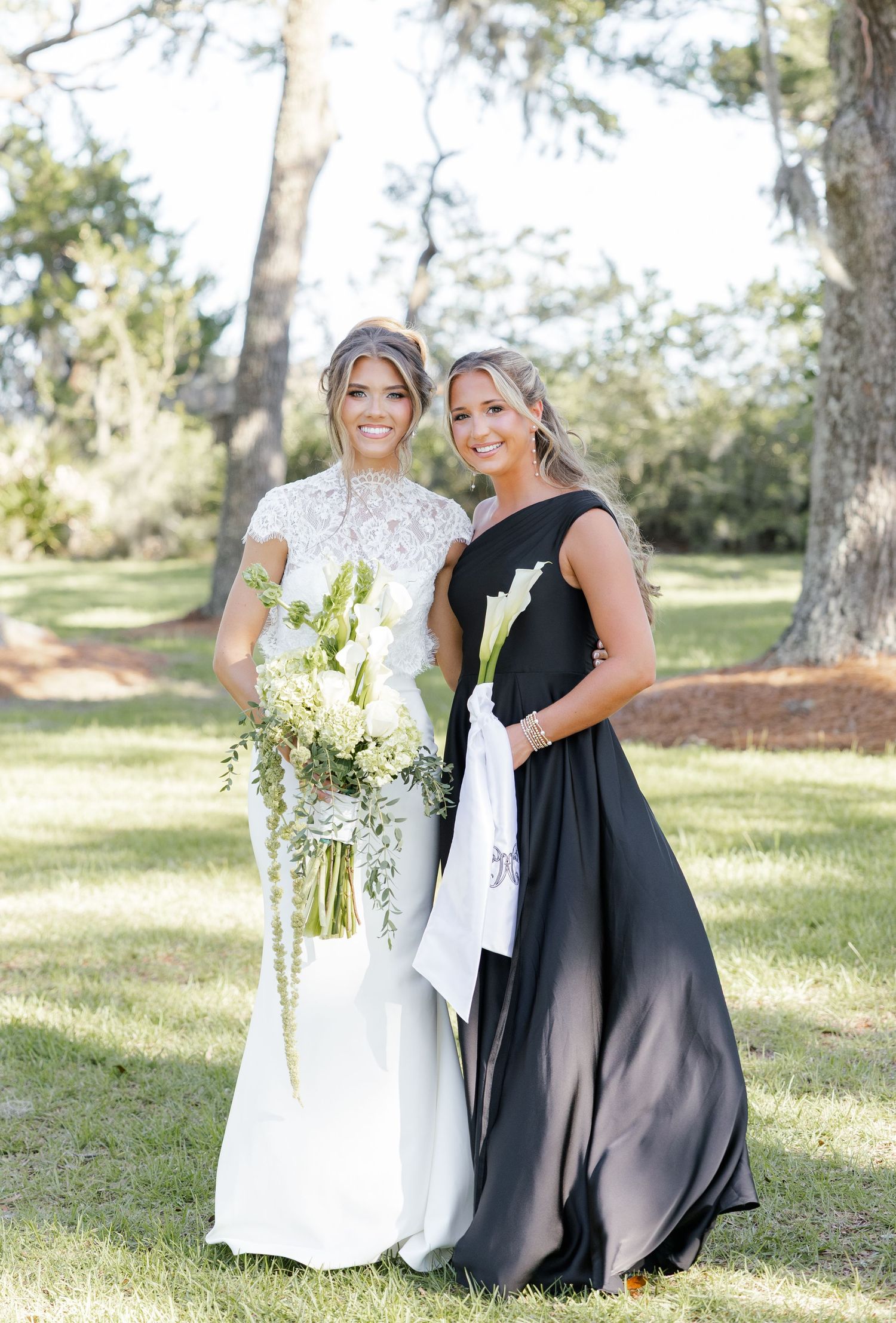 Bride in white lace gown holding bouquet stands with bridesmaid in black dress outdoors.
