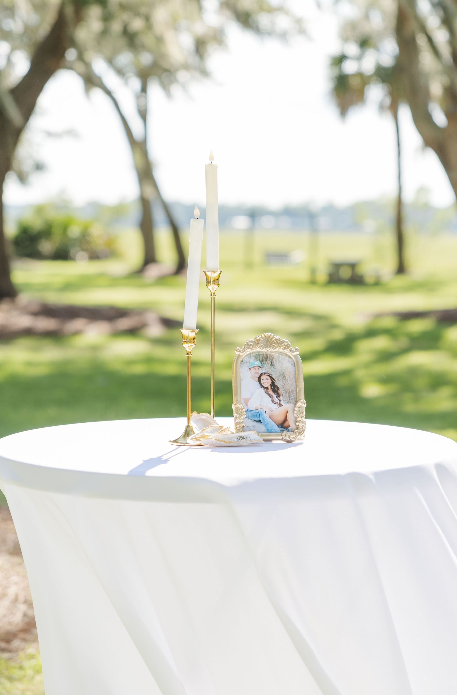 Elegant outdoor wedding memorial table with gold candlesticks and framed photo on white linen, set against lush green lawn.