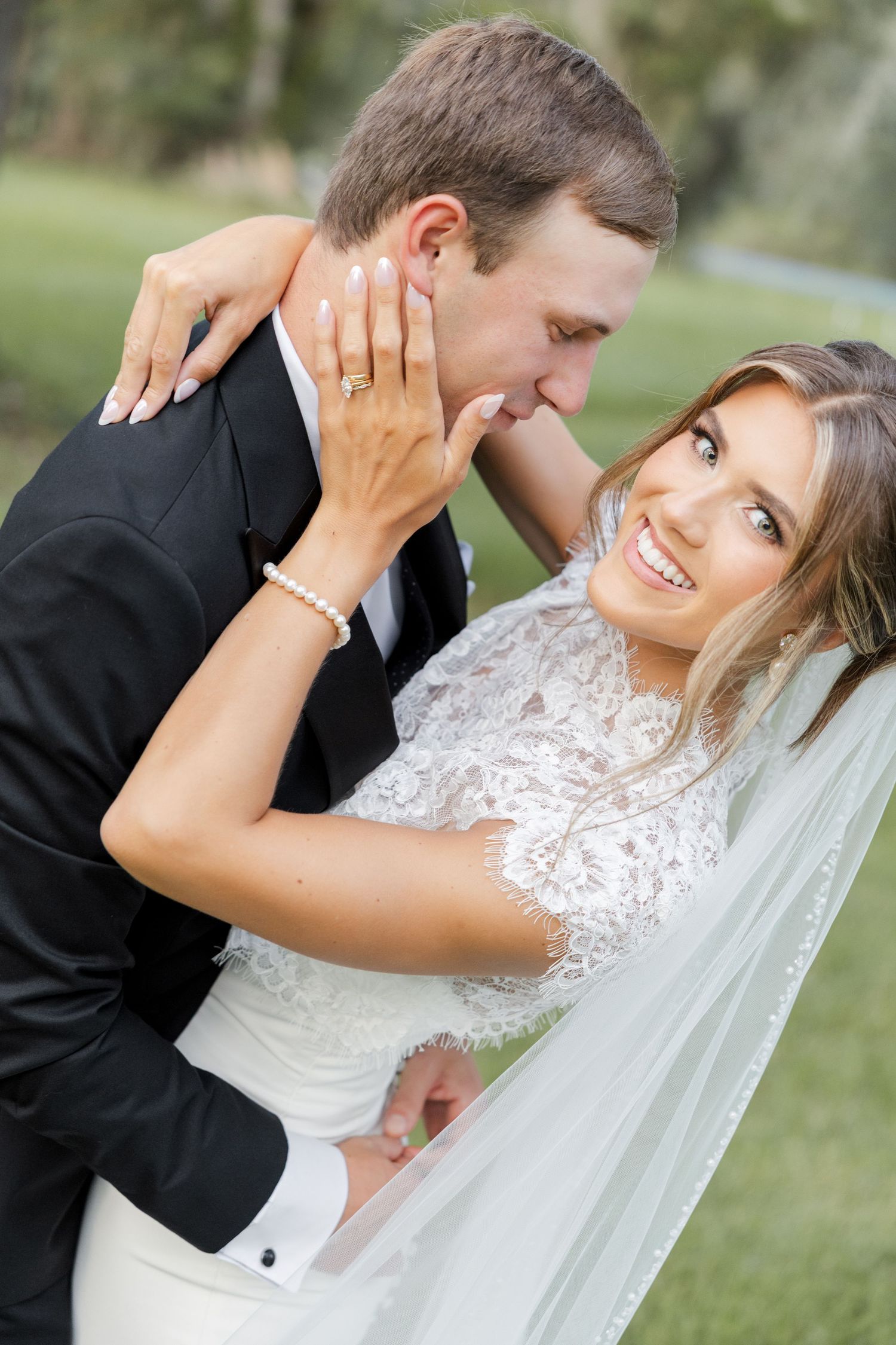 Bride in lace gown and veil smiles as groom in black tuxedo kisses her cheek outdoors.