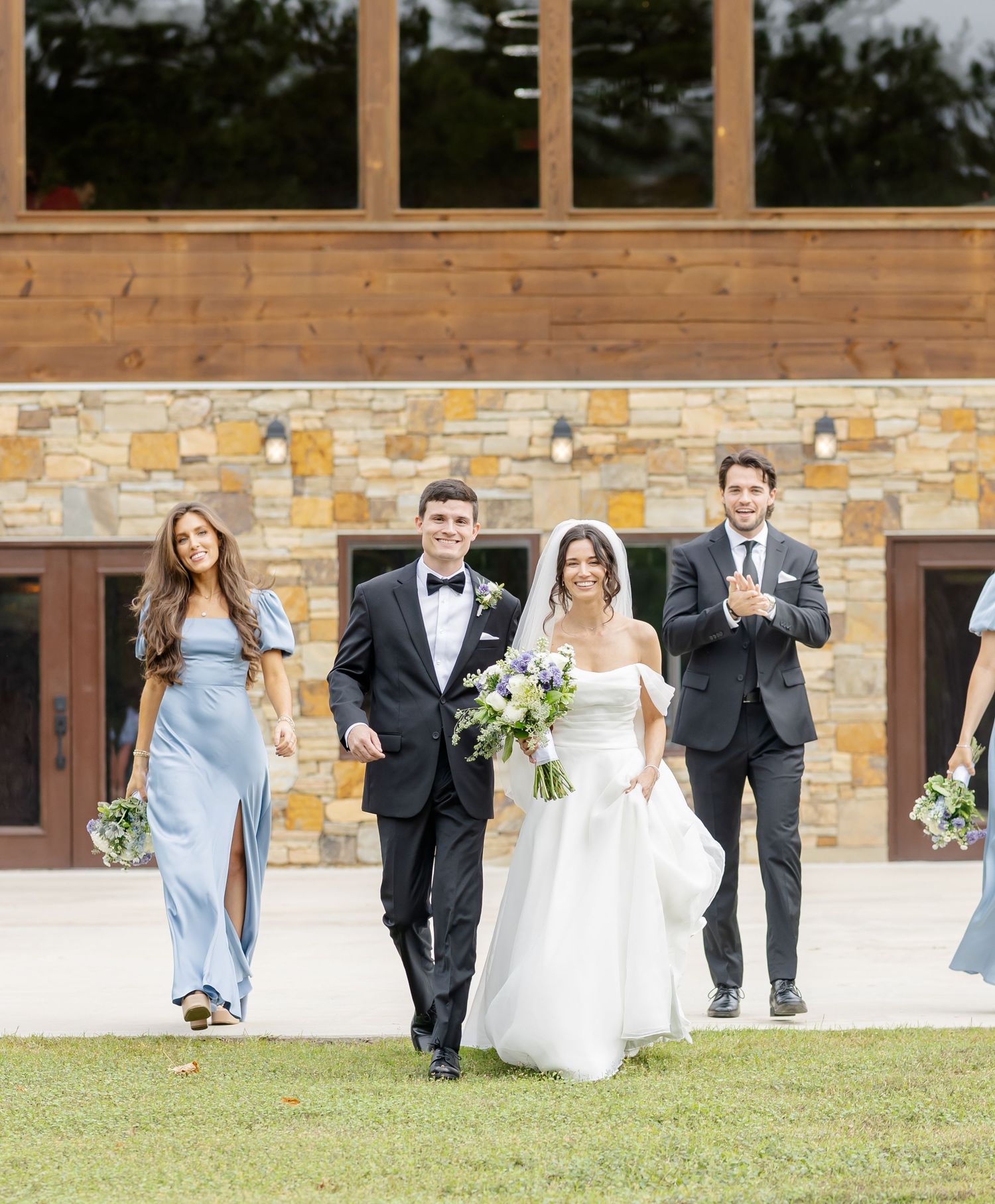 Wedding party in formal attire poses playfully on grassy lawn in front of stone building.