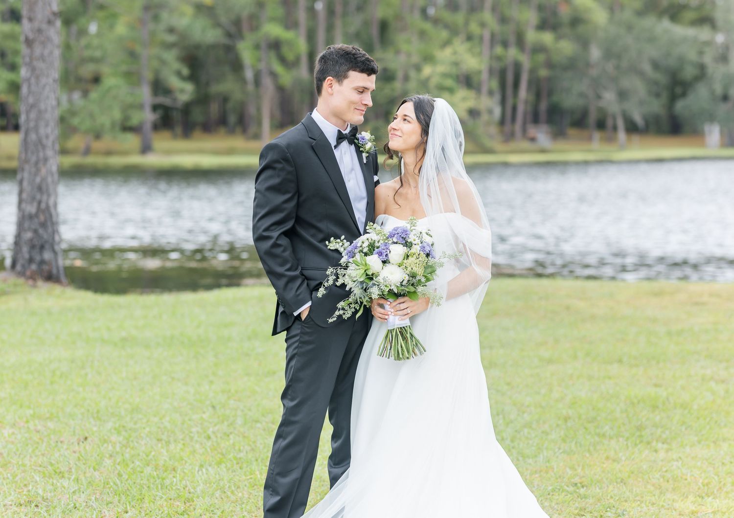 A couple in formal wedding attire shares a romantic moment outdoors by a serene lake with pine trees in the background.