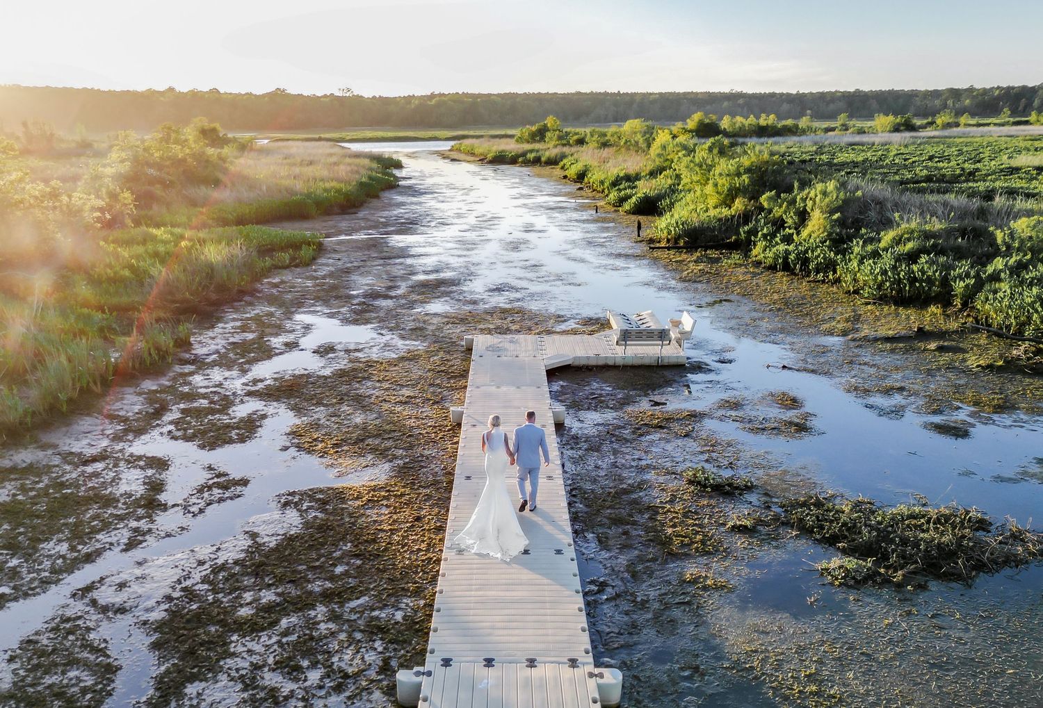 Aerial drone view of bride and groom walking on wooden dock through scenic coastal marshland at sunset.