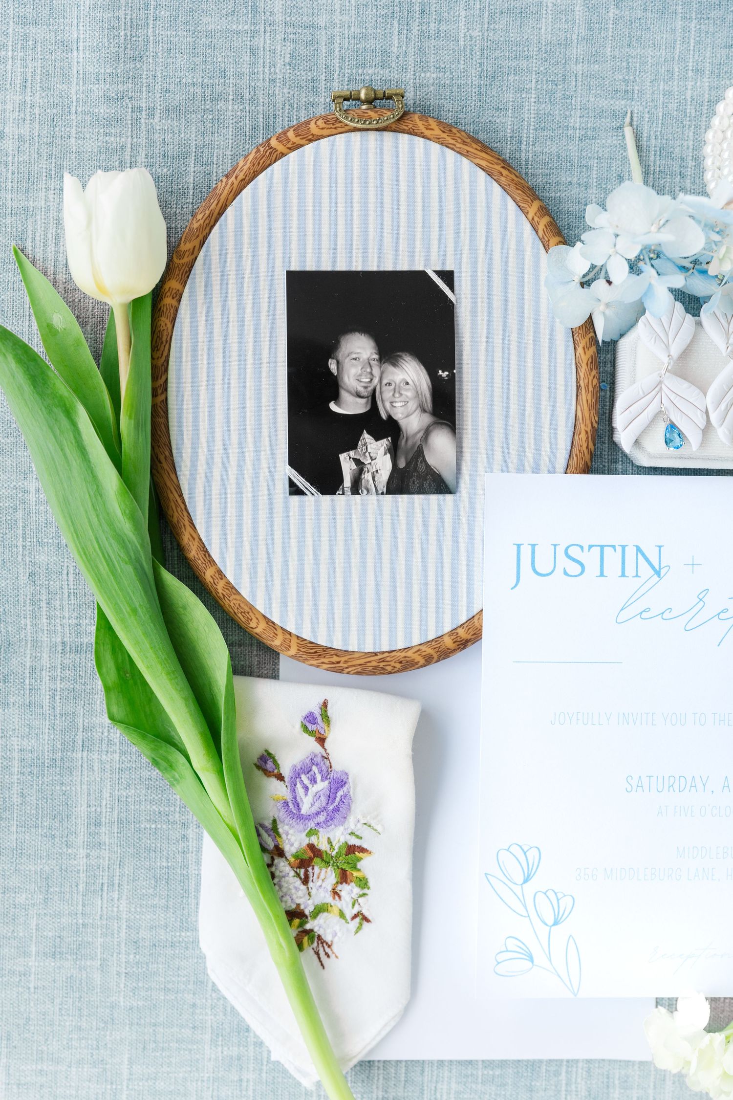 Blue and white striped embroidery hoop frame with black and white couple photo, white tulip, floral handkerchief, and wedding invitation.