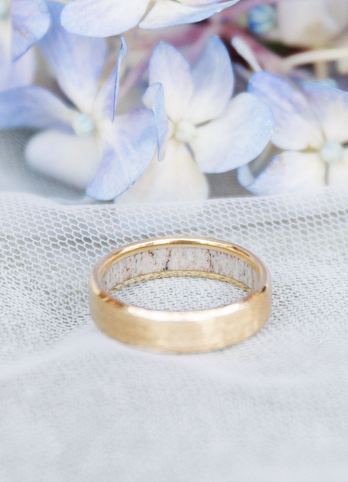 Gold wedding band with antler inlay displayed on white mesh fabric with soft blue hydrangea flowers in background.