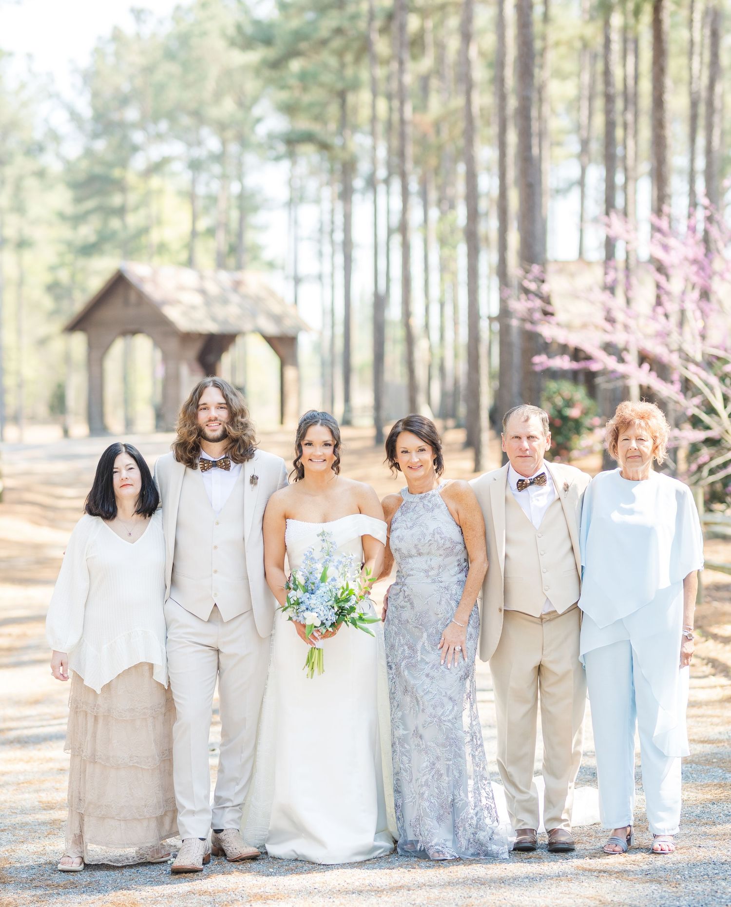 Wedding party family portrait outdoors among pine trees with blooming redbud tree in background.